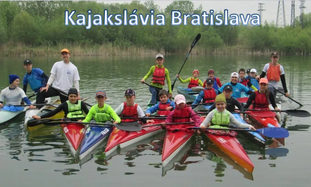 Group of children and teenagers in colorful life jackets sitting in kayaks on calm water with trees in the background and text 'Kajakslávia Bratislava' above.