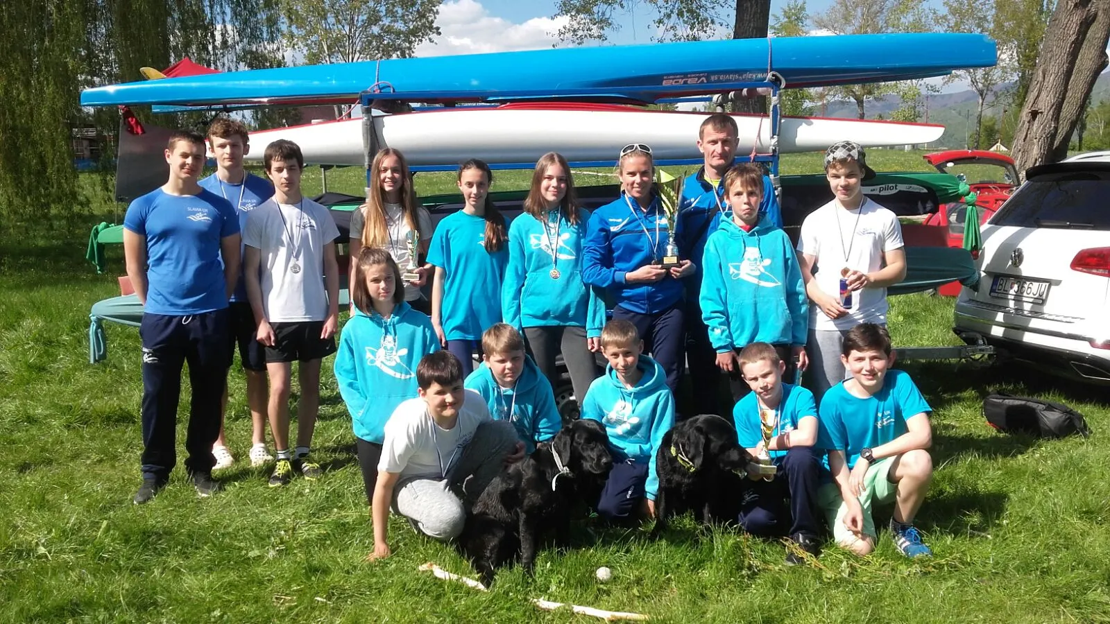 Group of teenagers and adults posing outdoors with medals, trophies, two black dogs, and kayaks on a rack in the background.