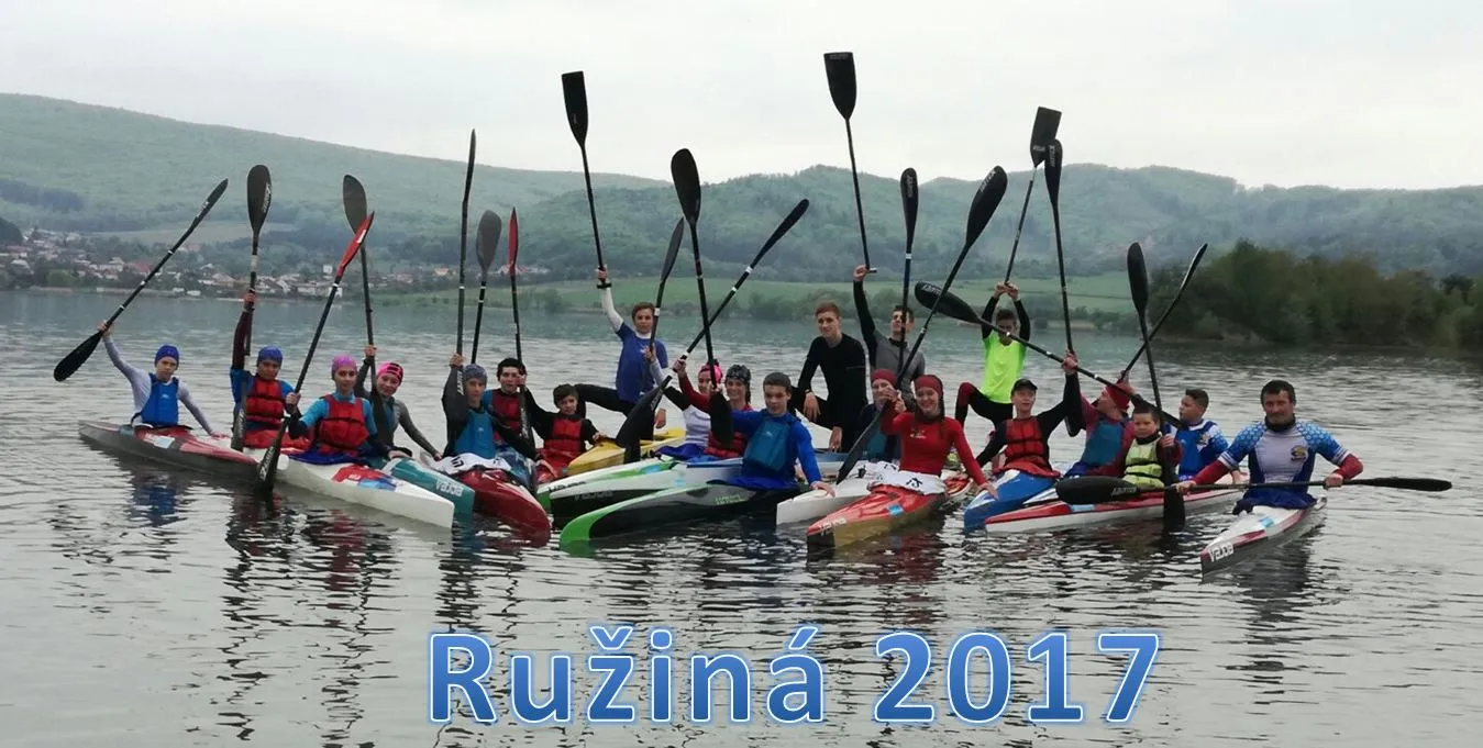 Group of kayakers on a lake holding paddles upright with hills and houses in the background.