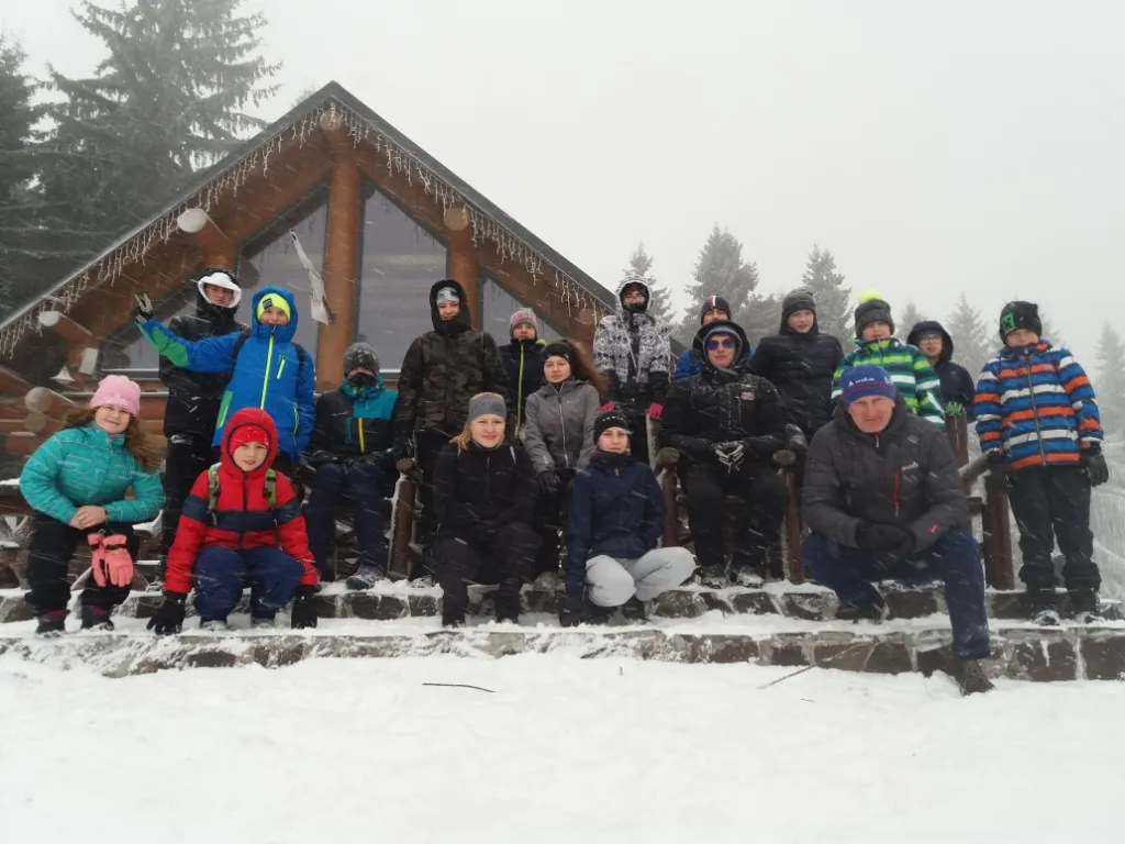 Group of people dressed in winter clothes posing on snowy steps in front of a wooden cabin with icicle lights.