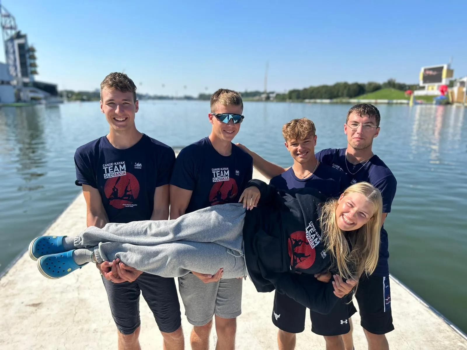Four young men smiling and holding a smiling young woman horizontally on a dock by a calm lake under a clear blue sky.
