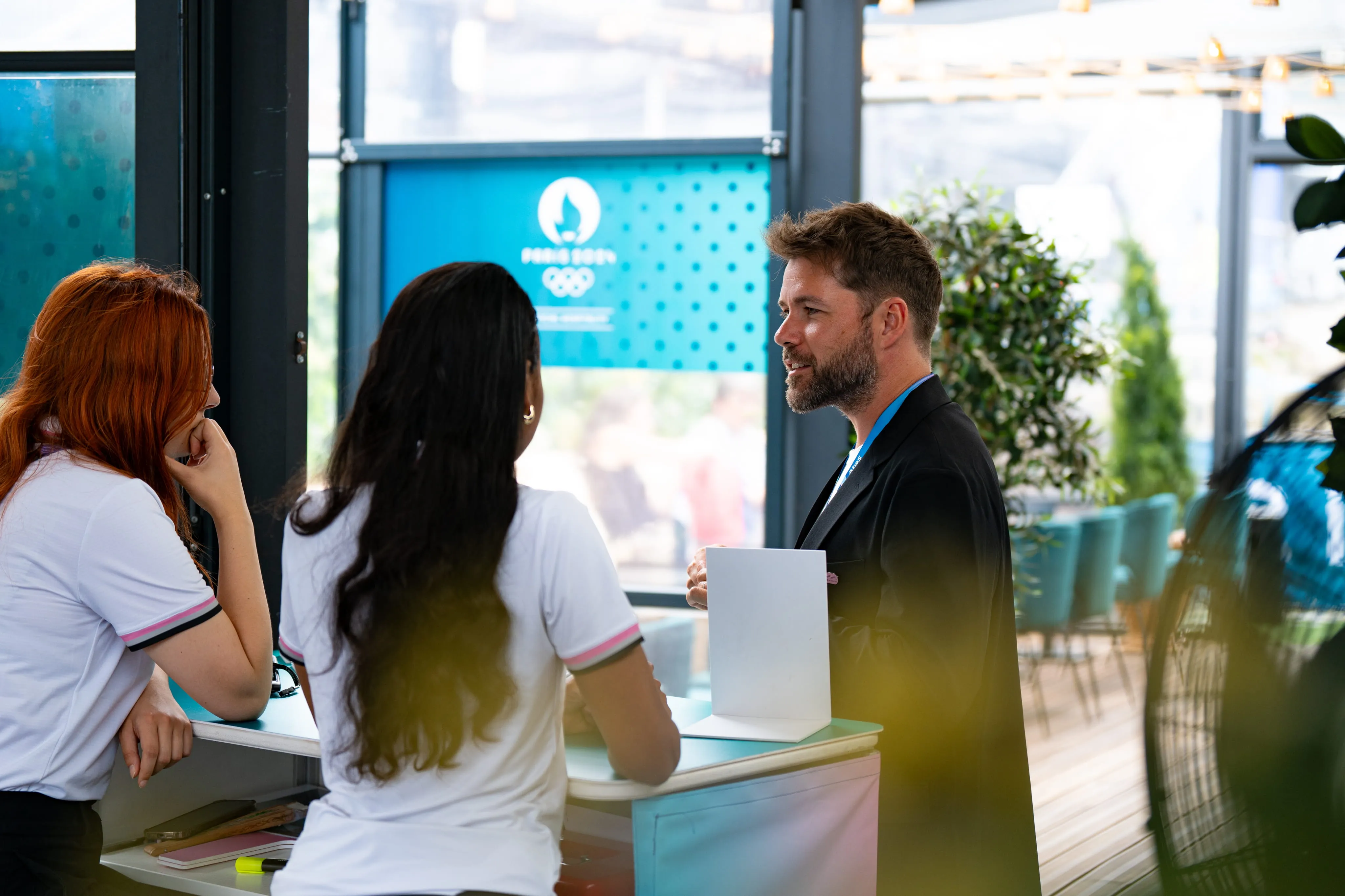 Man in a black blazer talking to two women in white shirts at a reception desk with a blue Paris 2024 Olympics sign in the background.