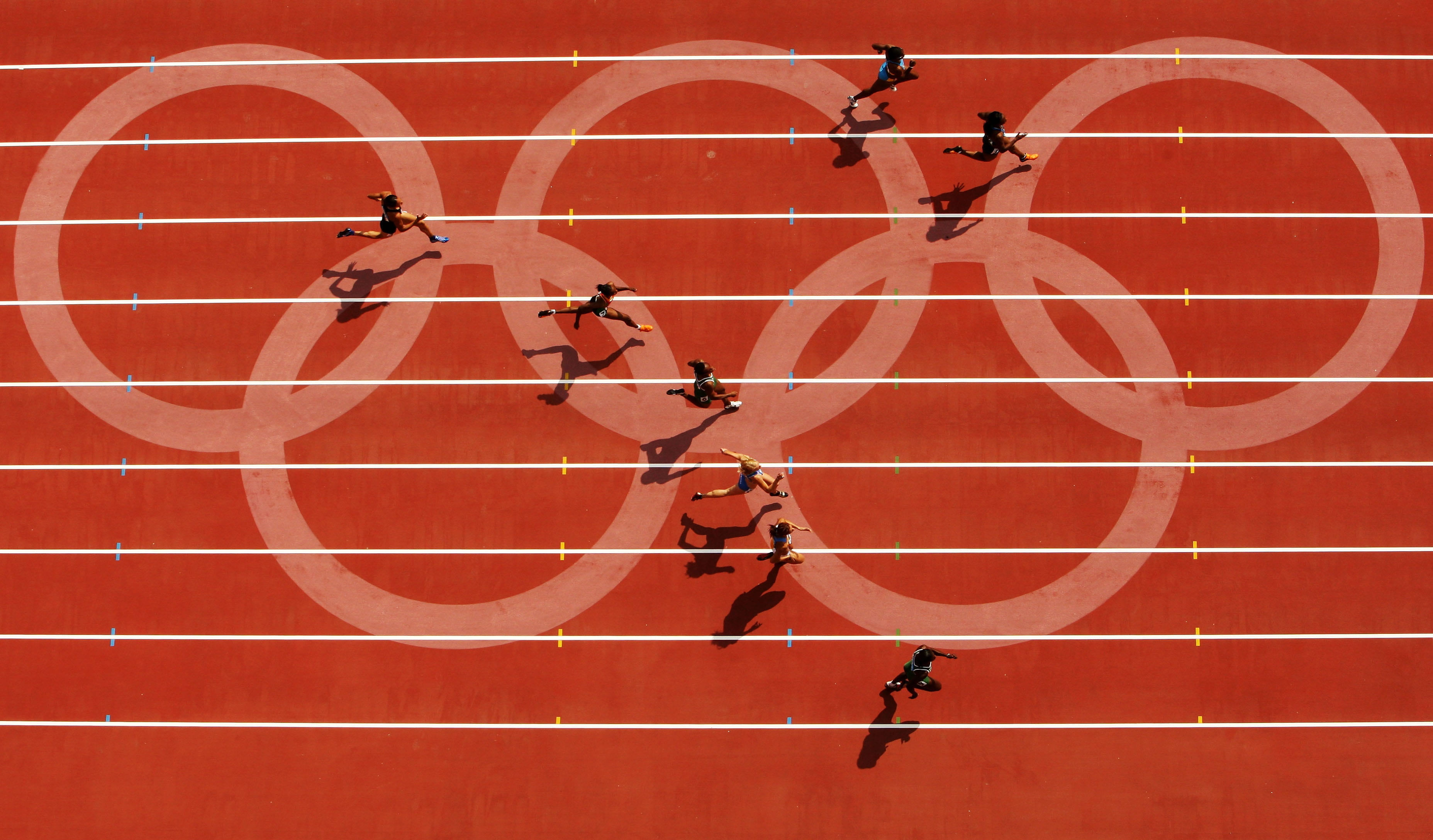 Overhead view of athletes sprinting on a red track with white lane markings and translucent Olympic rings in the background.
