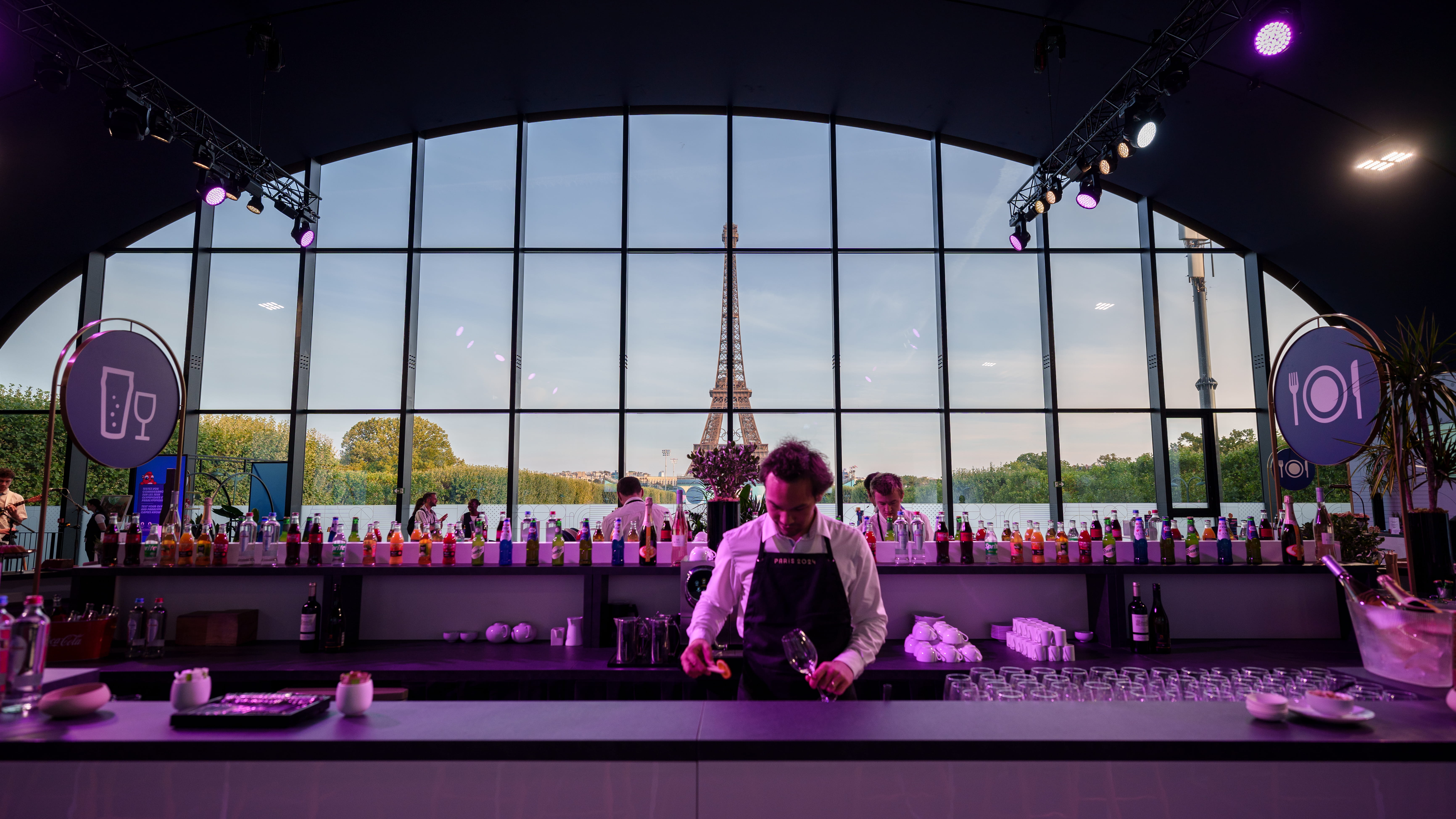 Bar inside a modern glass building with the Eiffel Tower visible outside through large windows, and a bartender preparing a drink.