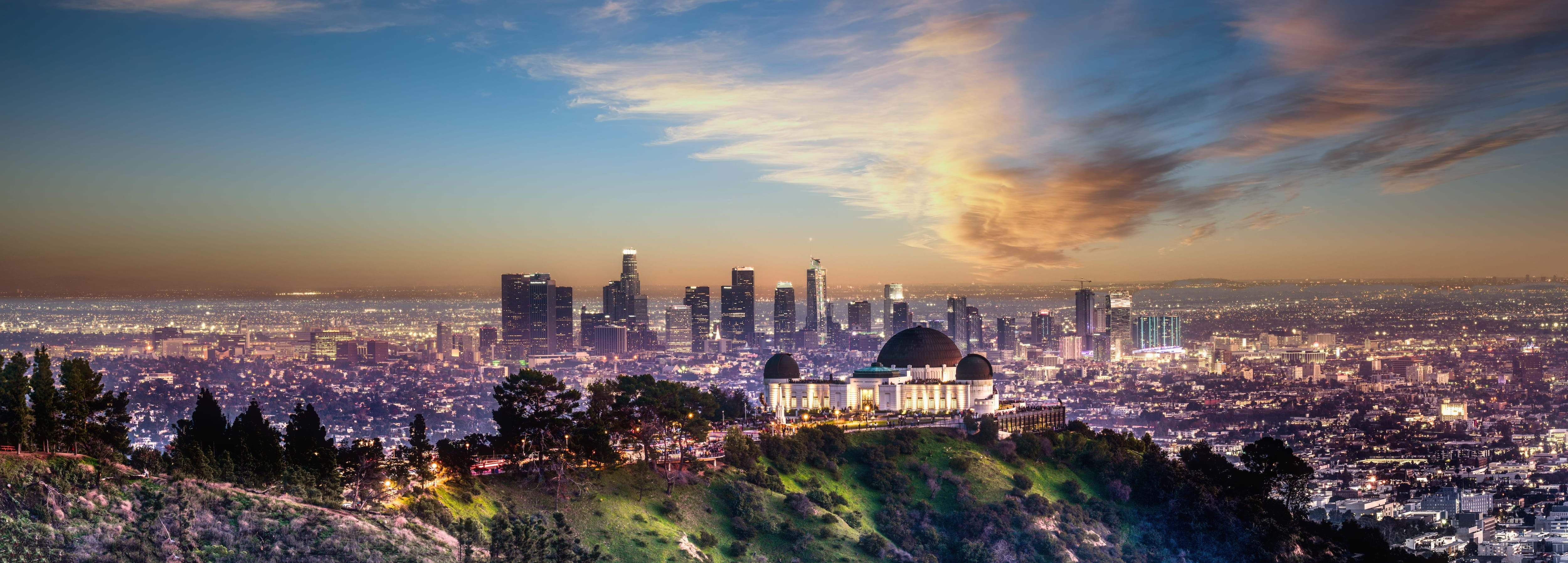 Panoramic dusk view of Los Angeles skyline with Griffith Observatory in the foreground on a green hill.