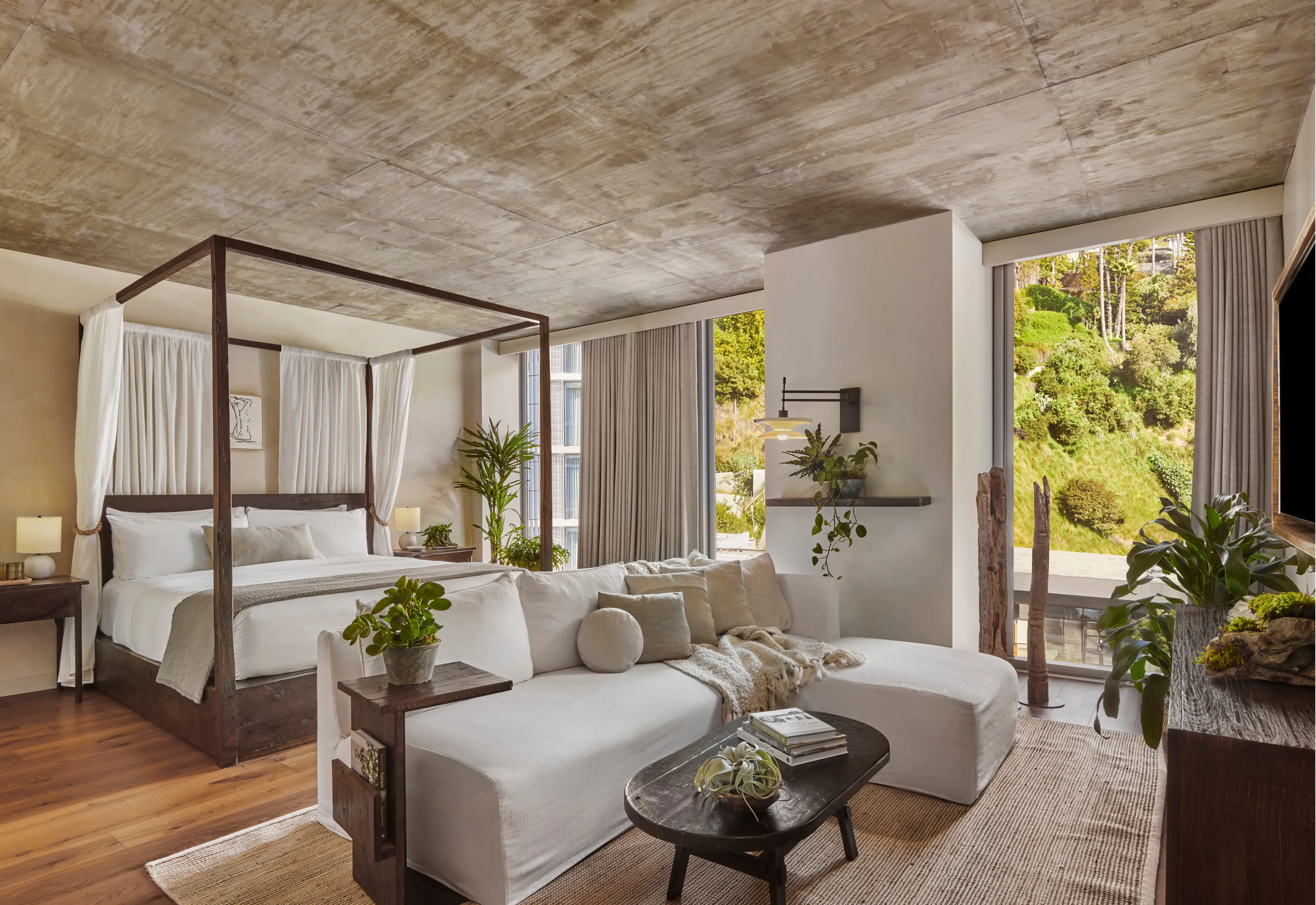 Modern bedroom with a wooden four-poster bed, white bedding, a white sectional sofa with cushions and throw, wooden coffee table and large windows overlooking greenery.