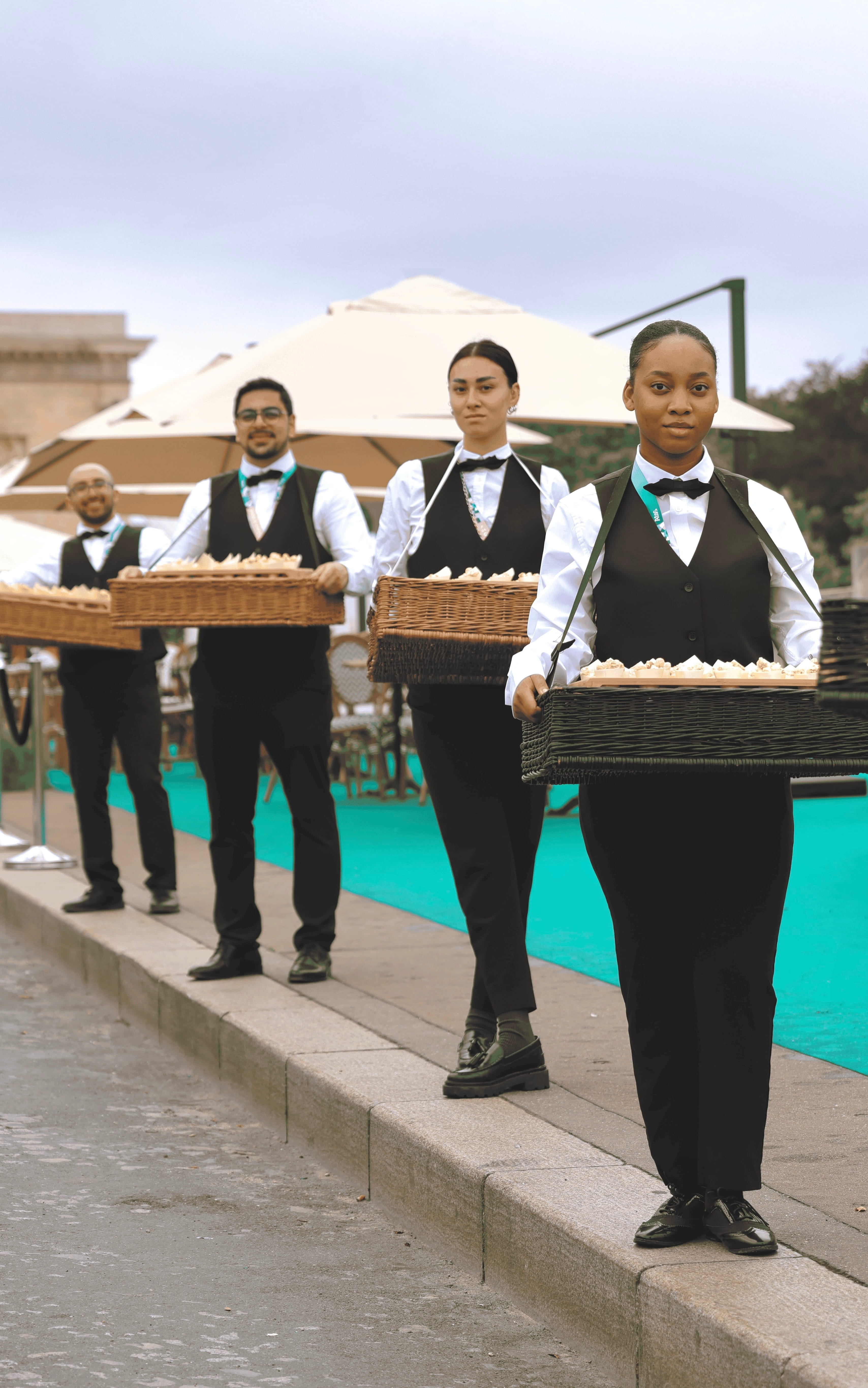 Four servers dressed in black vests, white shirts, and bow ties carrying trays of appetizers along a street near outdoor umbrellas.