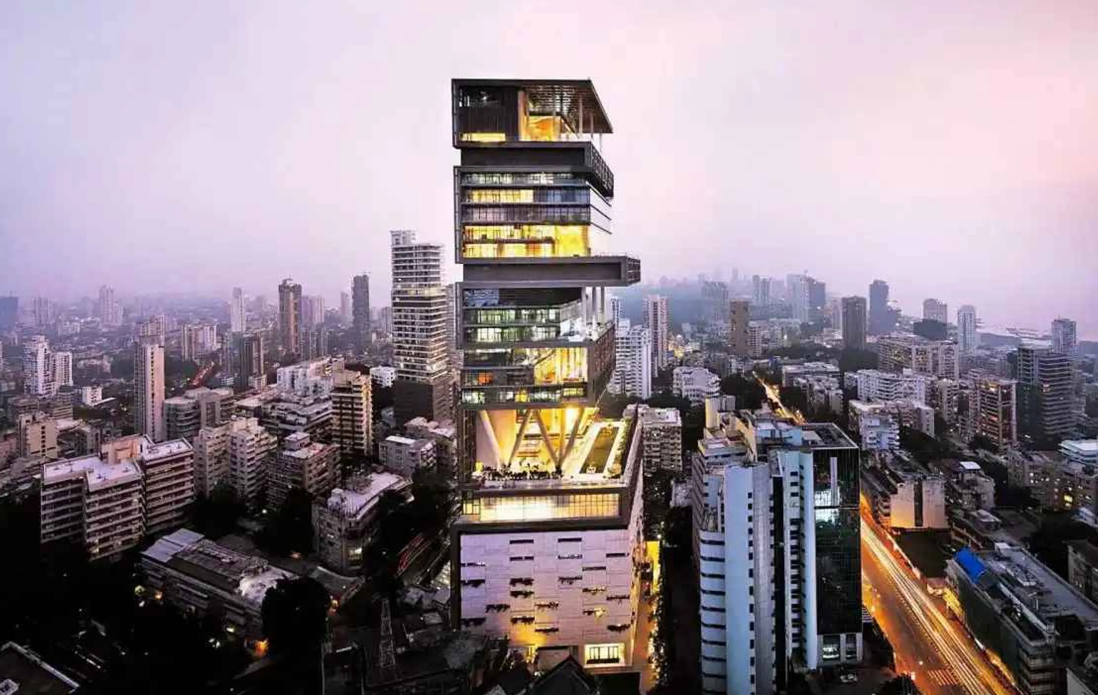The illuminated Antilia skyscraper with a unique cantilevered design amidst a dense urban cityscape at dusk.