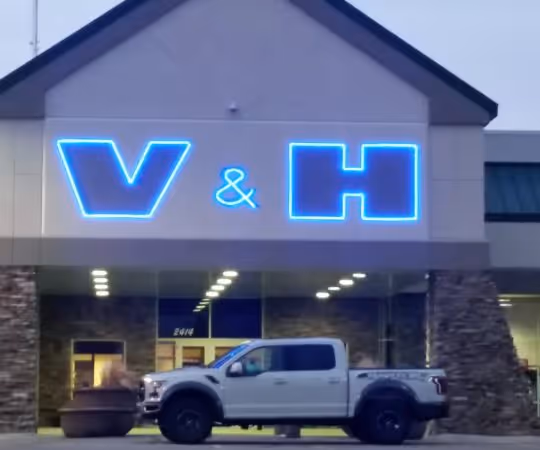 White pickup truck parked in front of a building with a glowing blue neon V & H sign above the entrance.