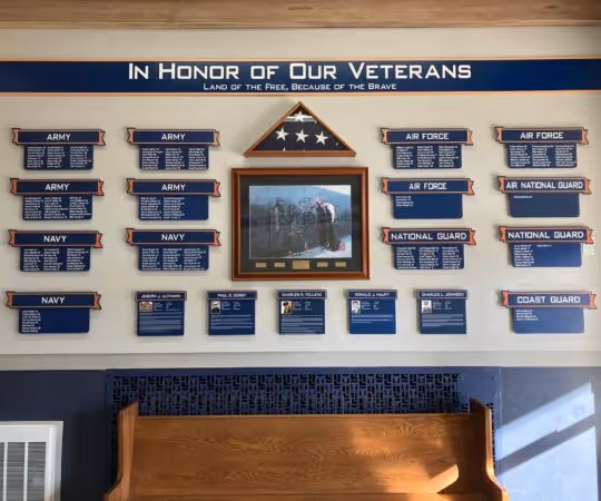 Memorial wall titled 'In Honor of Our Veterans' with plaques for Army, Navy, Air Force, Air National Guard, National Guard, and Coast Guard, a folded American flag, and a framed photo above a wooden pew.