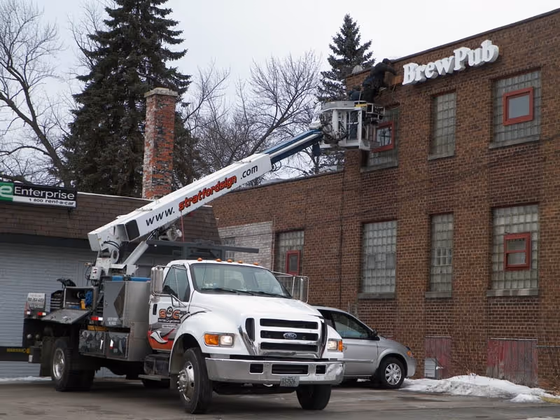 White utility truck with an elevated work platform where a worker is installing or repairing a BrewPub sign on a brick building.