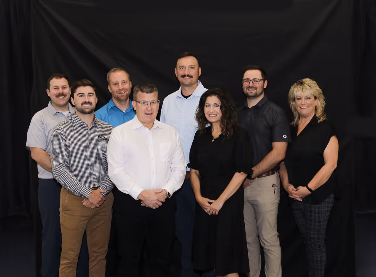Group portrait of five men and three women standing in two rows against a black backdrop, all smiling and dressed in business casual attire.