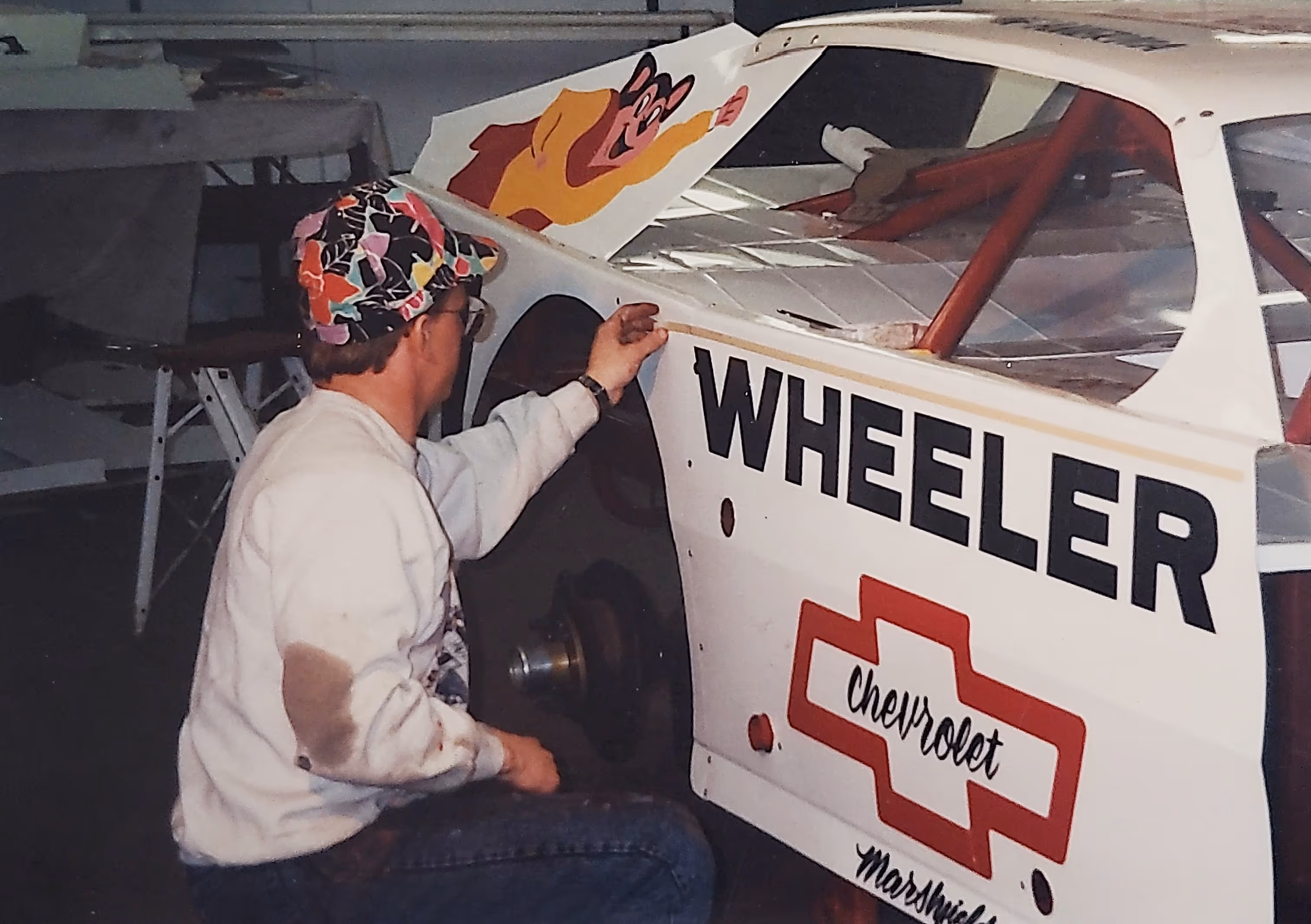 Man in a colorful cap and white sweatshirt working on the wheel area of a white racing car with WHEELER and Chevrolet logos.