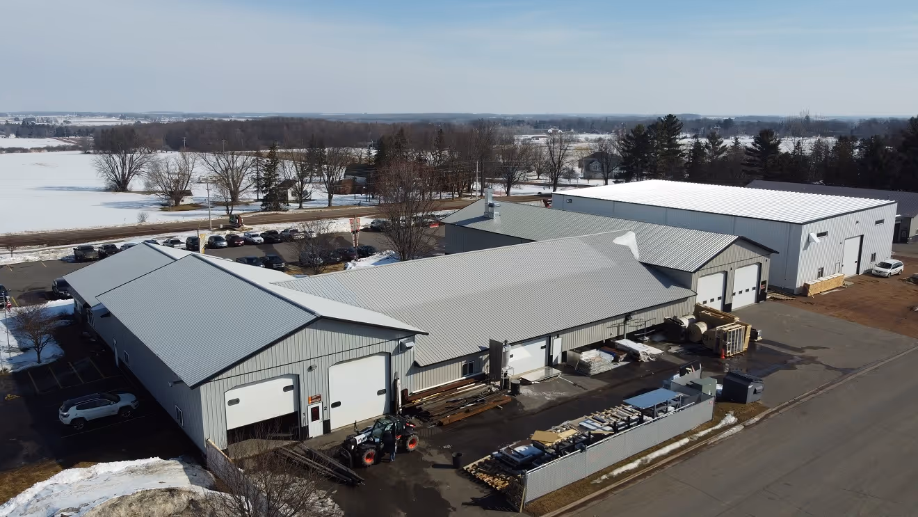 Aerial view of industrial warehouse buildings with metal roofs surrounded by parked vehicles and snowy landscape.