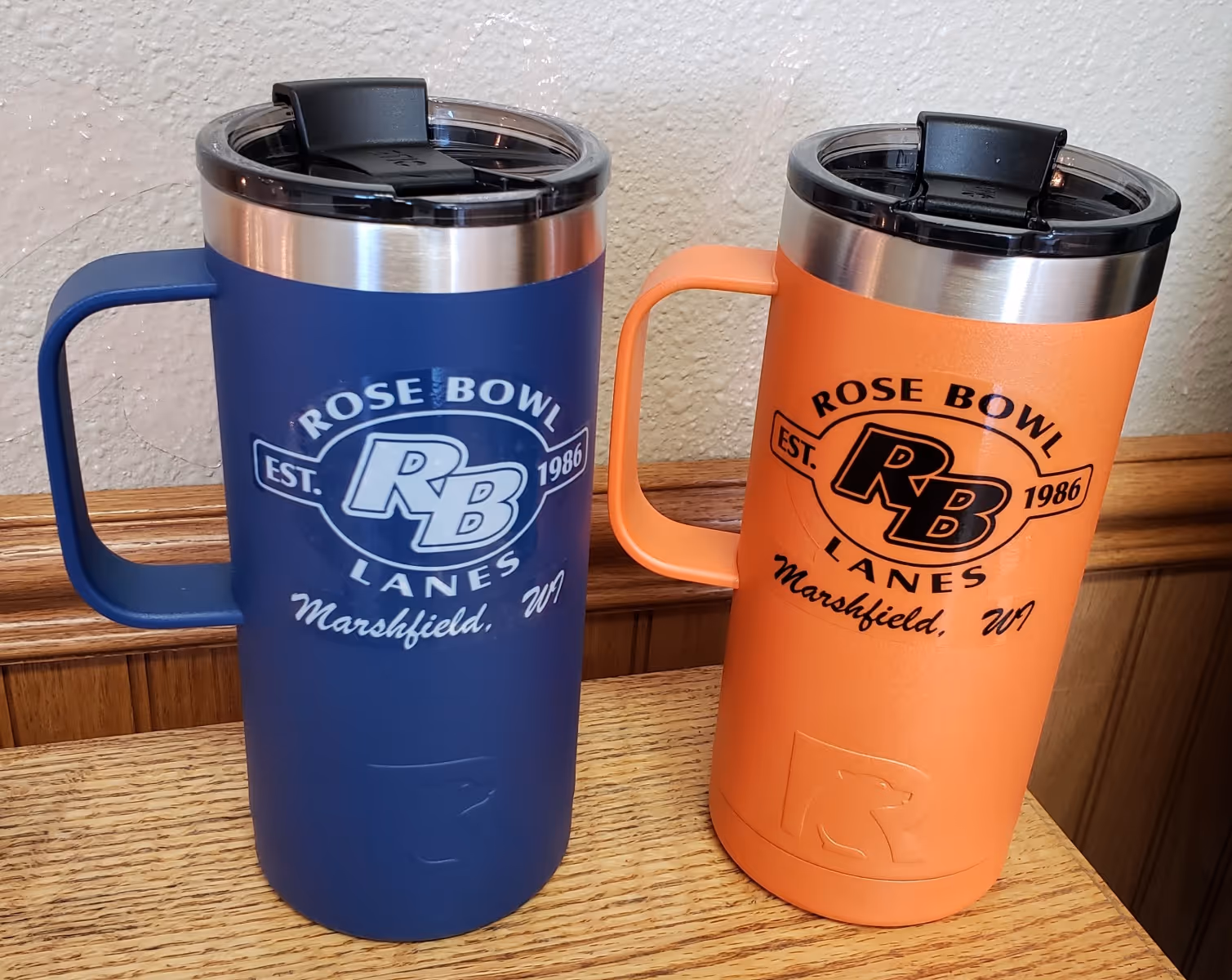 Blue and orange travel mugs with black lids, each featuring the Rose Bowl Lanes logo and text 'EST. 1986 Marshfield, WI,' placed on a wooden surface.