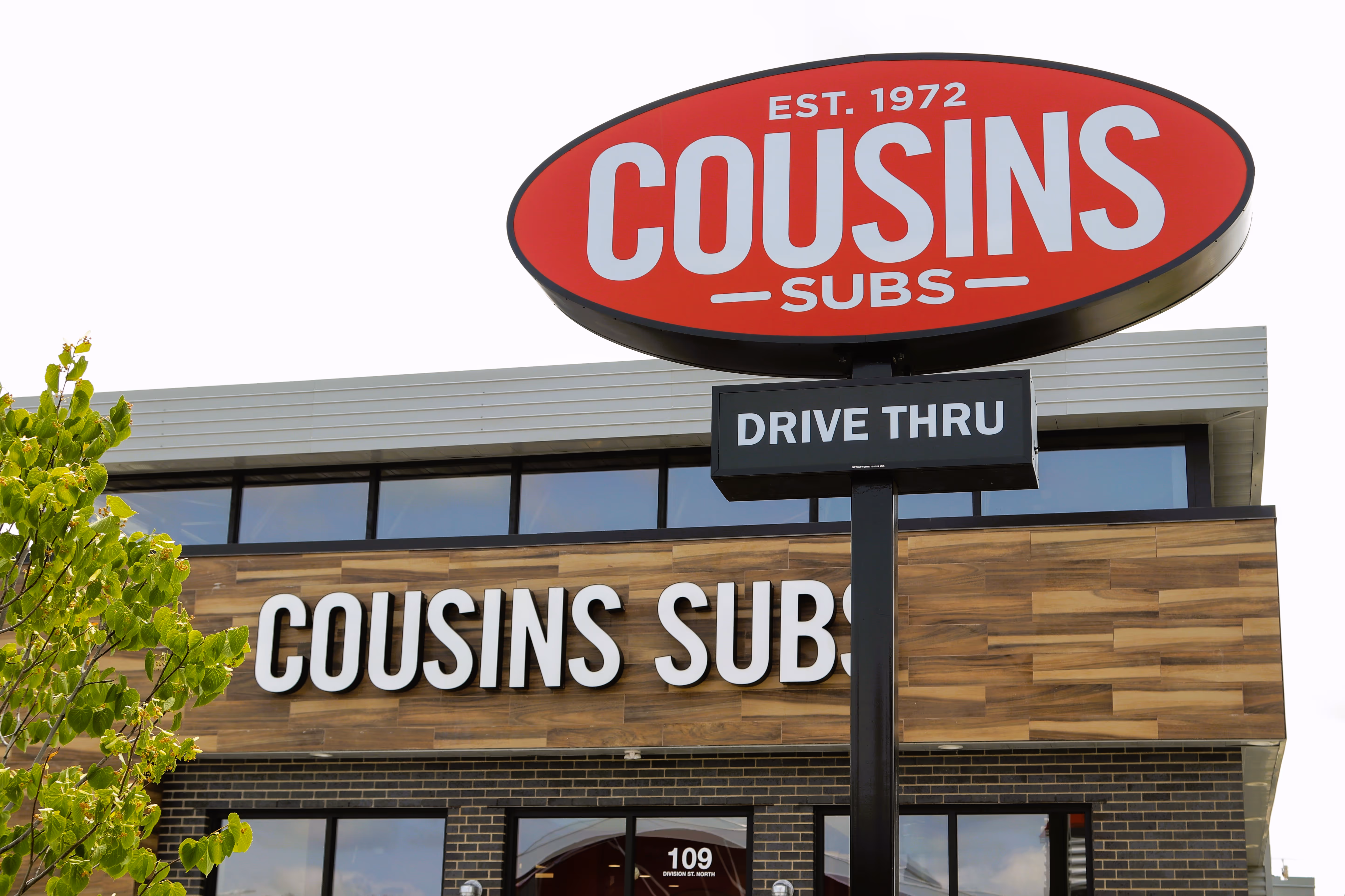 Cousins Subs restaurant exterior with a red oval sign and a drive-thru sign in front.