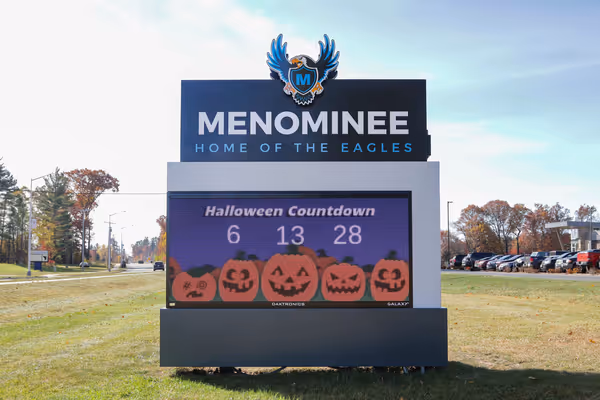 Outdoor digital sign for Menominee school with a Halloween countdown showing 6 days, 13 hours, and 28 minutes, decorated with jack-o'-lantern graphics.