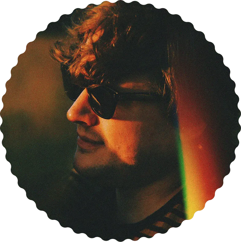 Side profile of a young man with curly hair wearing sunglasses and lit by warm, colorful light.