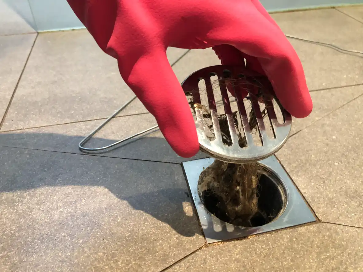 A hand in a red glove holds a metal drain strainer full of hair and grime, pulling it from a floor drain.