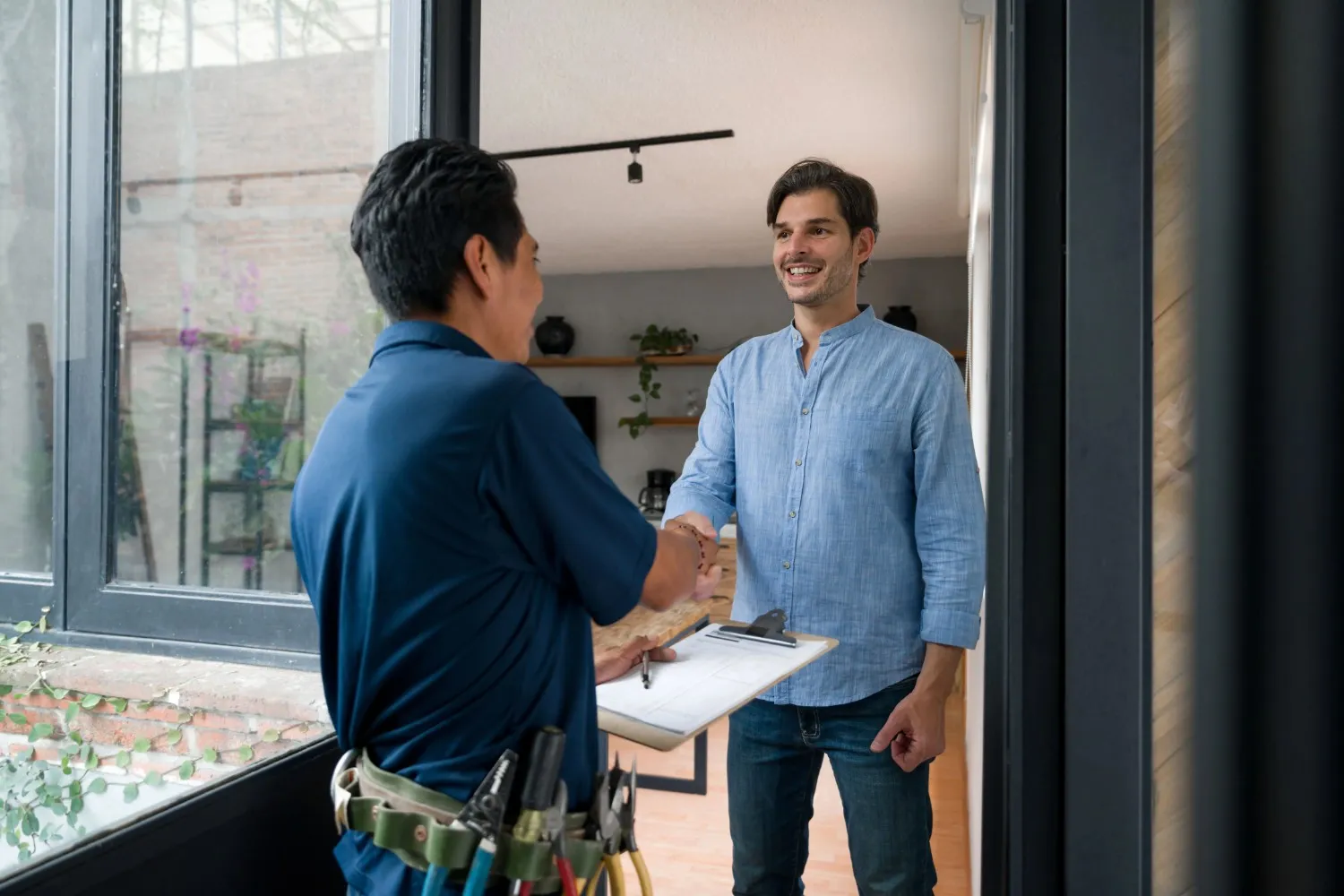 A smiling man in a light blue shirt shakes hands with an electrician holding a clipboard.