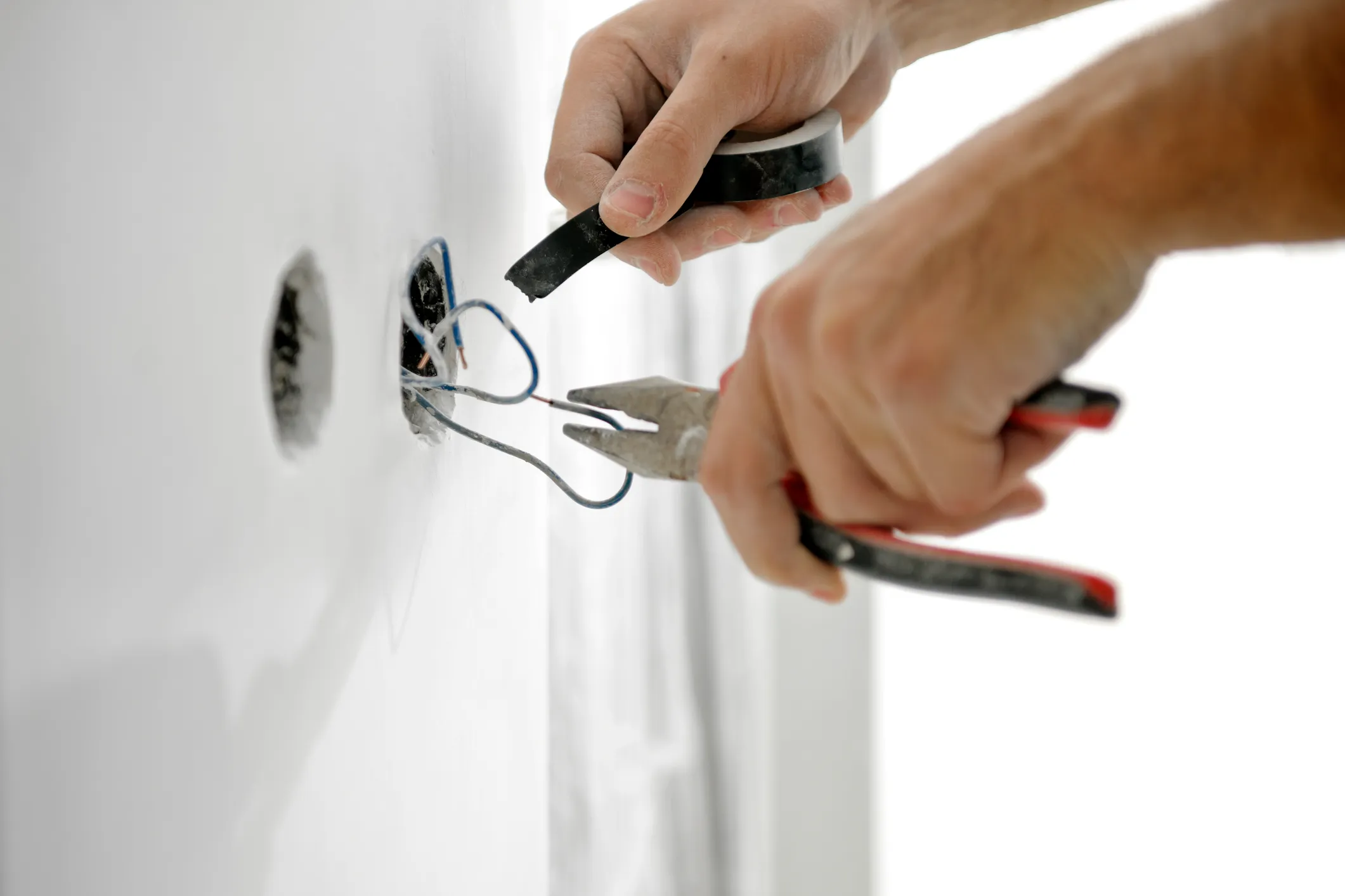 An electrician's hands use pliers and electrical tape to work with wires coming out of a wall hole.