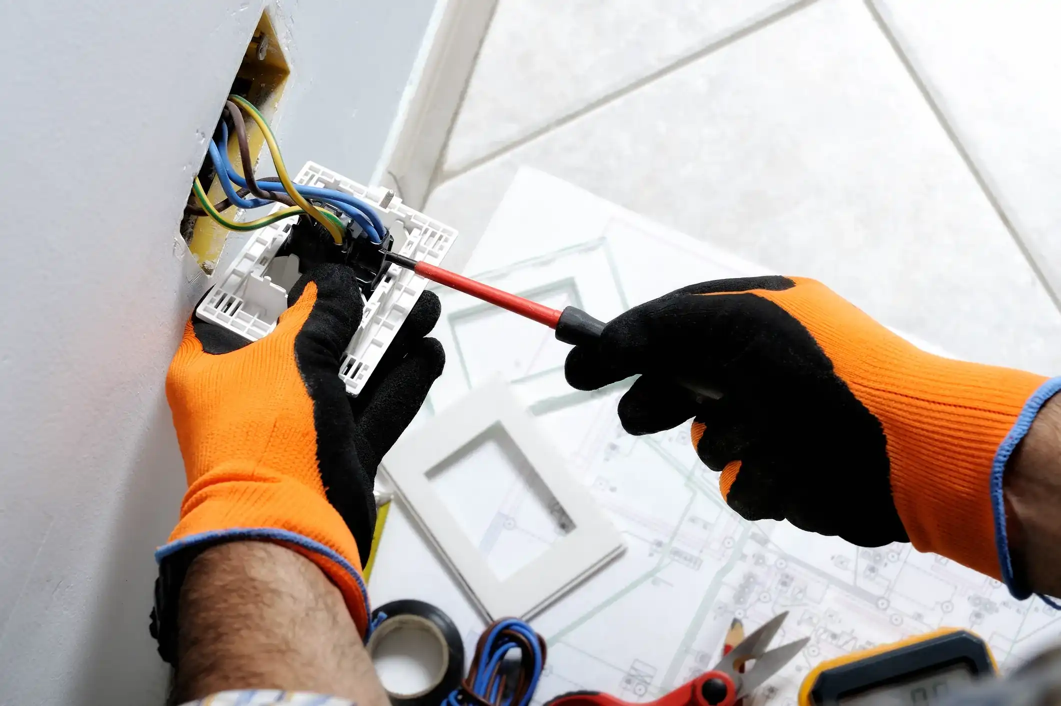 An electrician with orange and black gloves uses a screwdriver to connect wires to an electrical switch plate.