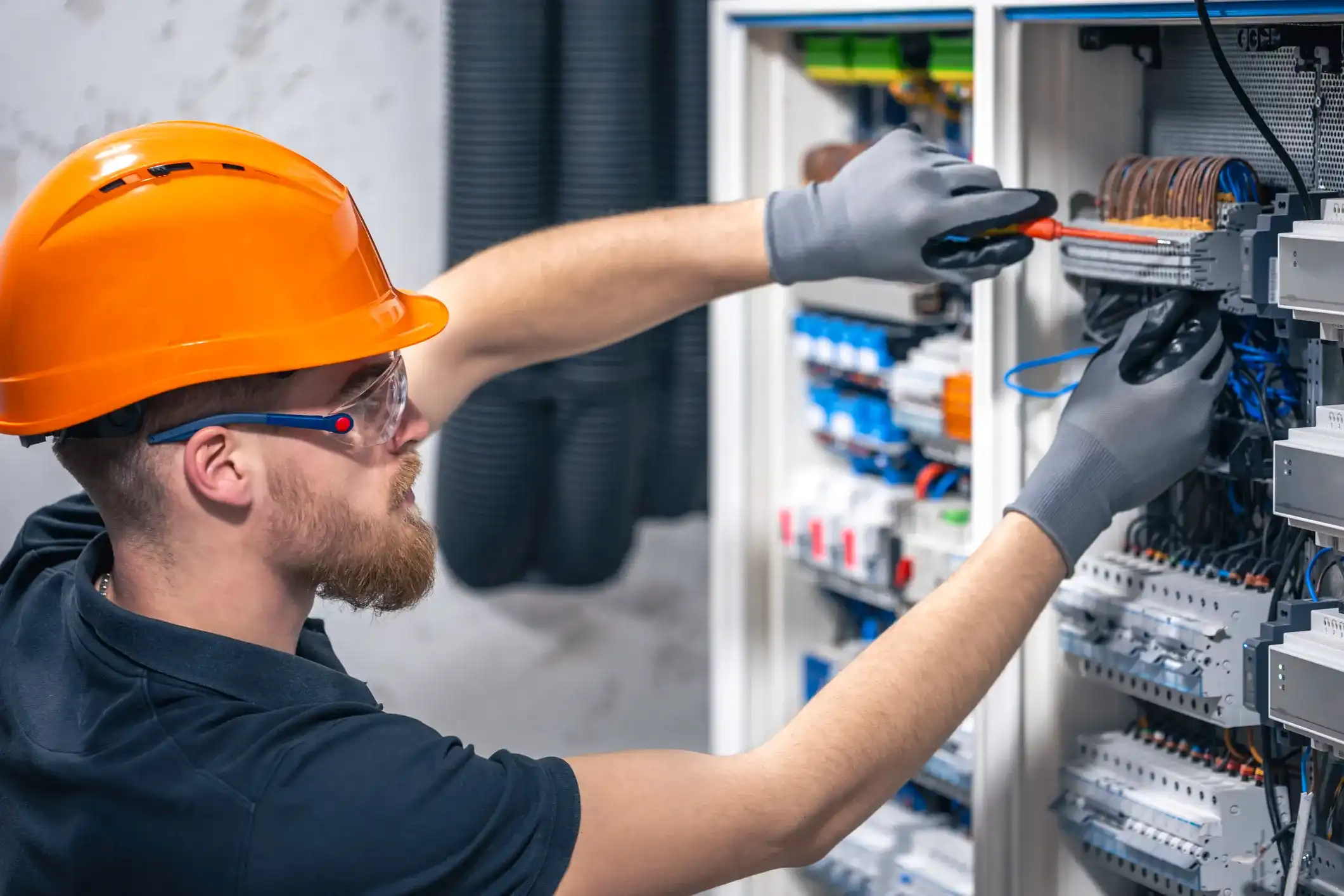 A focused electrician with a beard, wearing an orange hard hat and safety glasses, uses a screwdriver to work on a circuit board in a breaker box.