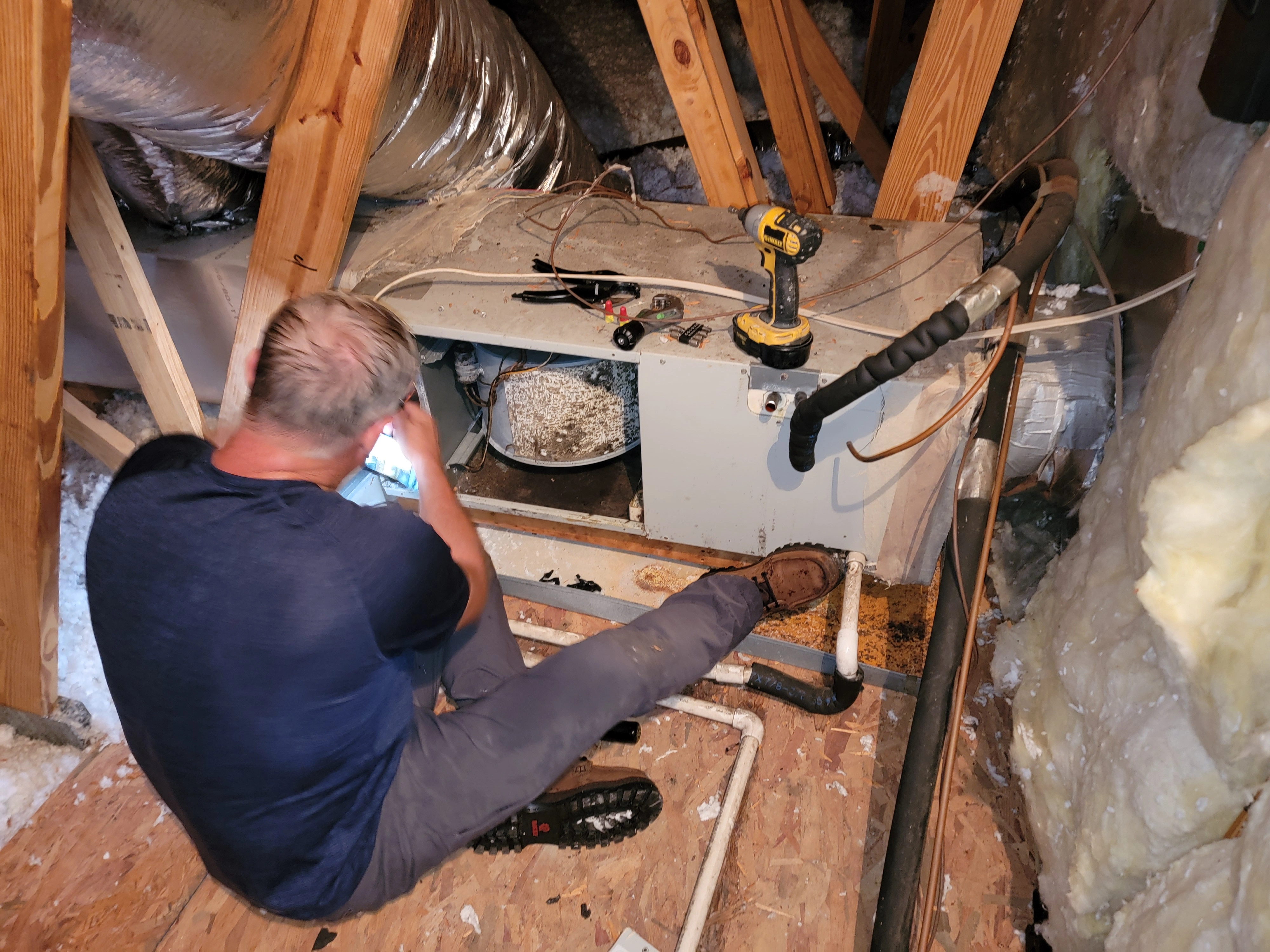 Man working on attic HVAC unit, surrounded by insulation and wooden beams. 