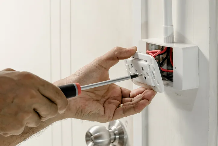 A person's hands are shown using a screwdriver to install a new electrical outlet.