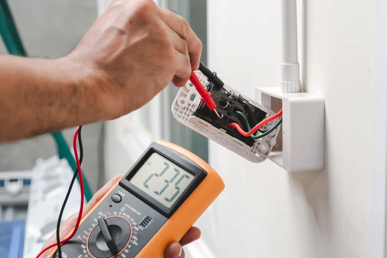 A close-up of a hand using a multimeter to test an electrical outlet with a bare wire.