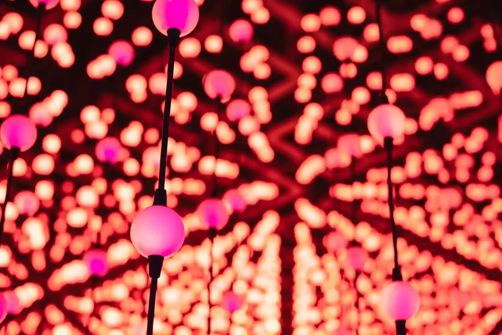 A close-up, abstract image of glowing pink spherical lights hanging from black cords against a background of blurred, red bokeh.