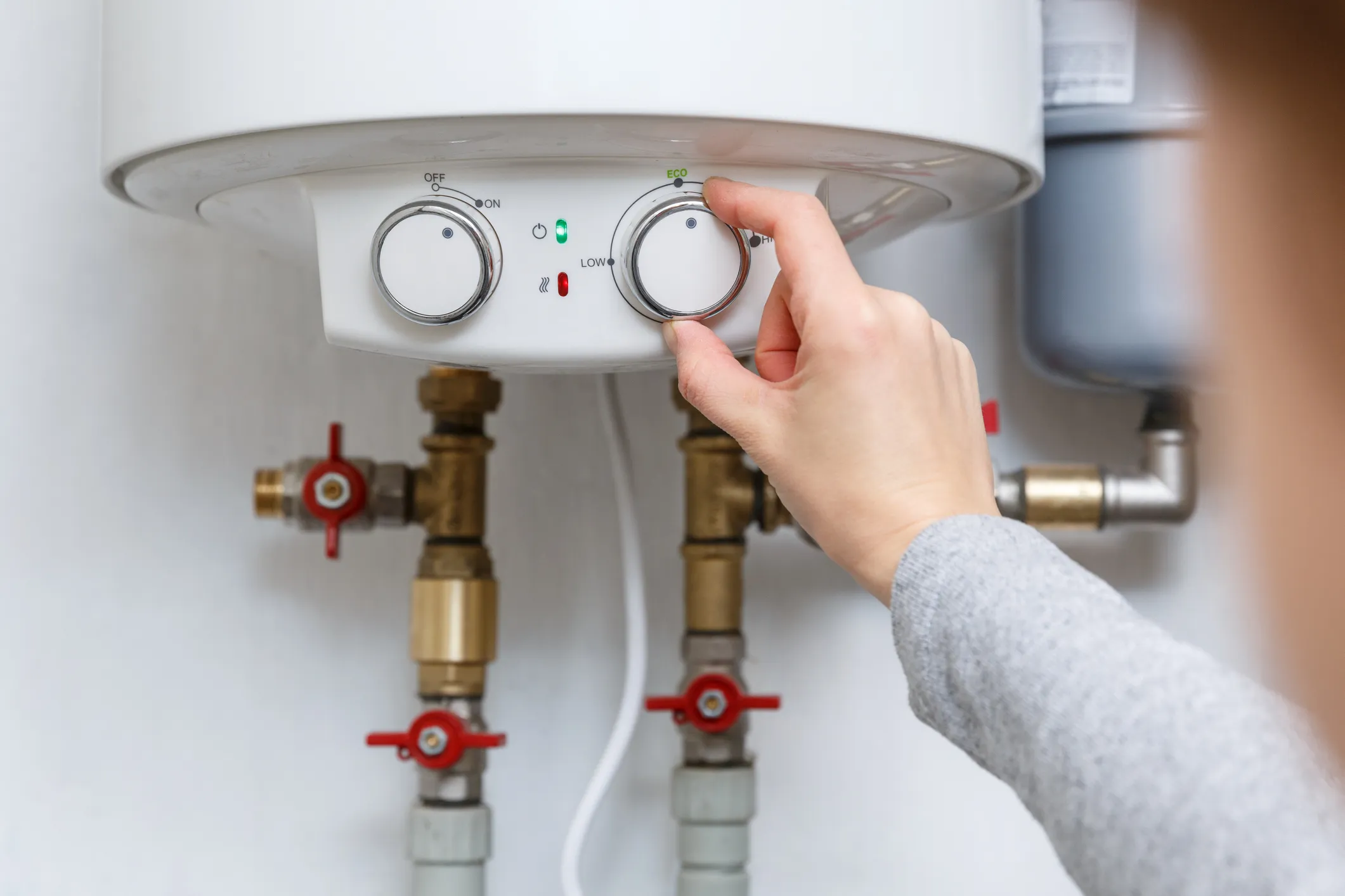 A person's hand is adjusting a dial on the control panel of a white, tankless water heater, which shows settings for "Off," "On," and "Eco."