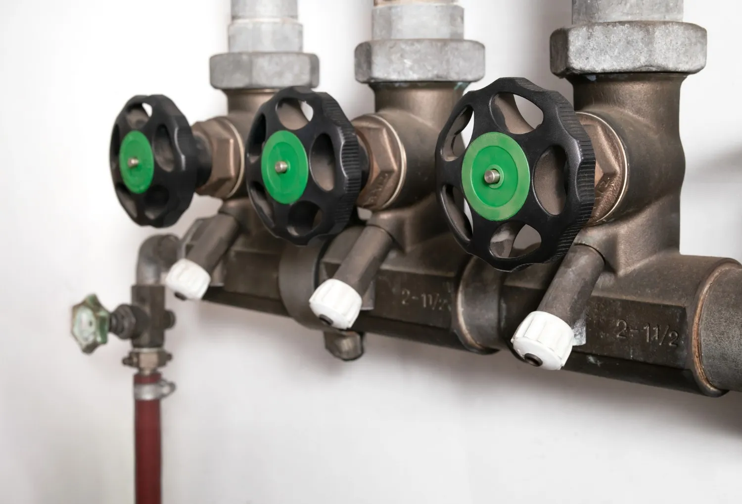 A close-up of a plumbing backflow prevention valve with three black and green handles and a series of pipes mounted against a white wall.