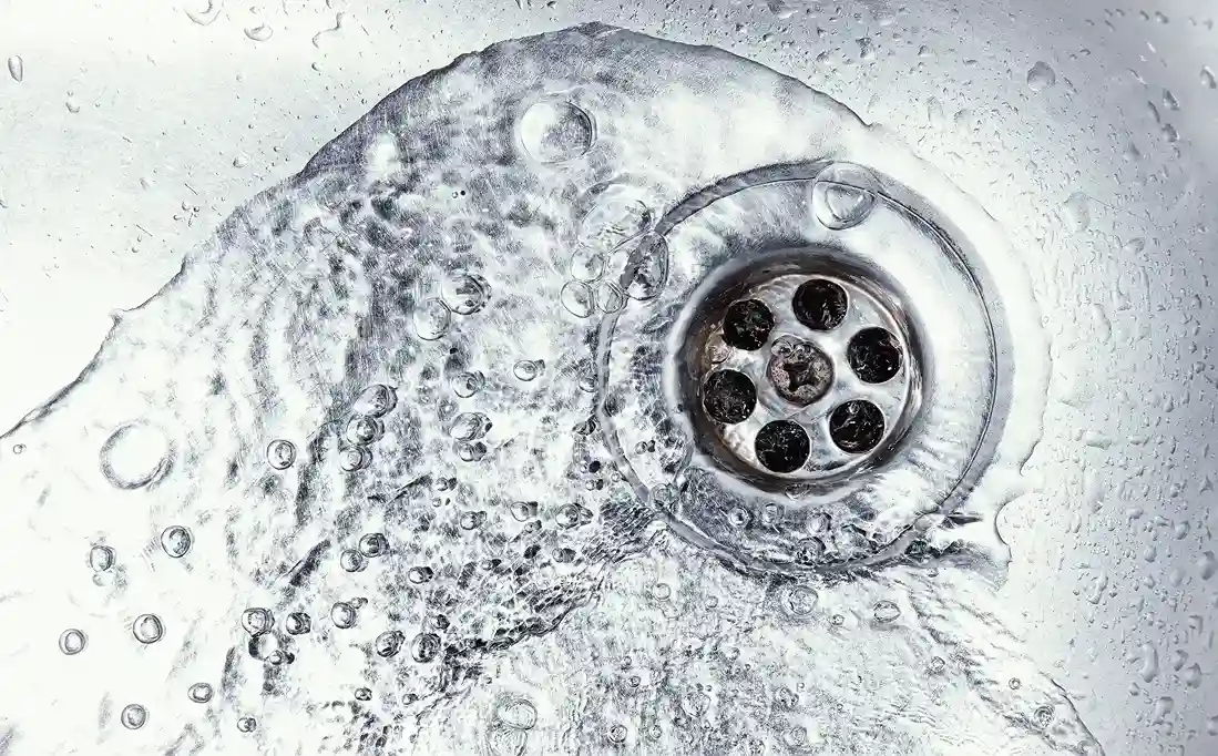 A close-up, overhead view of a clean, shiny sink drain as clear water with bubbles swirls around it and flows downward.