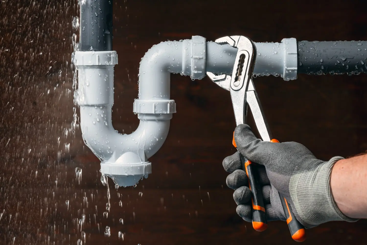 A plumber's gloved hand holds a wrench to a leaking white plastic drain pipe, with water spraying and splashing against a dark wooden background.