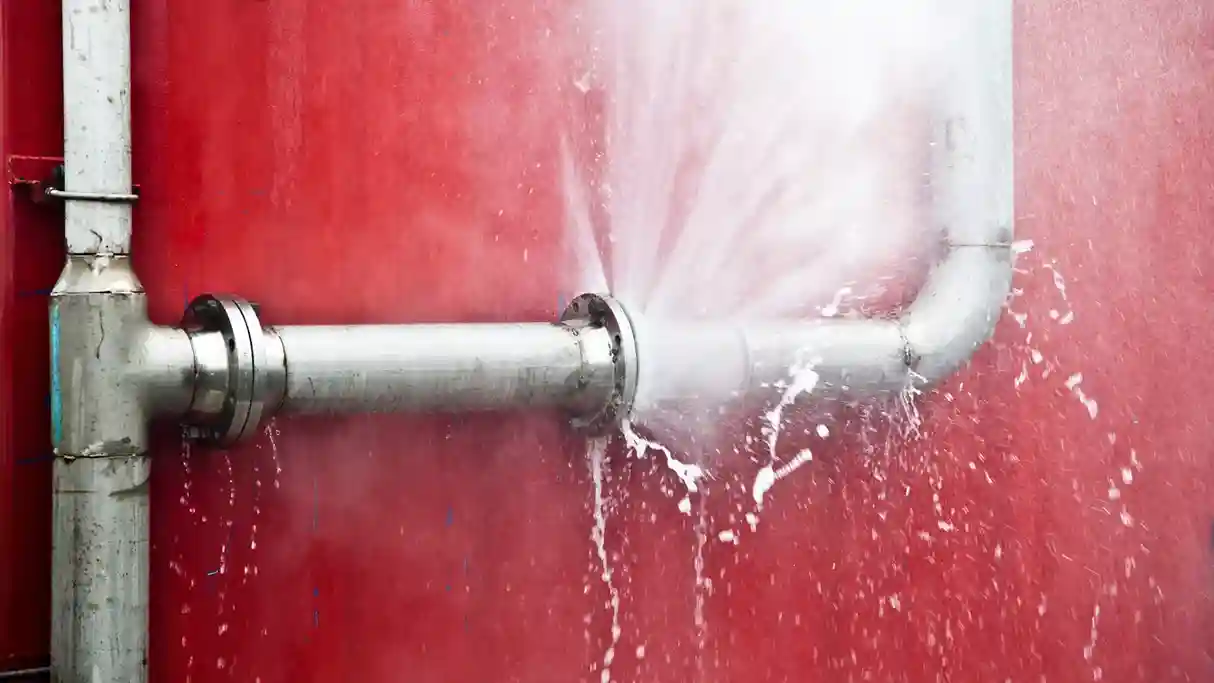 A close-up shot of a severe leak from a silver metal pipe with a flange connection, spraying a strong jet of water against a red industrial wall.