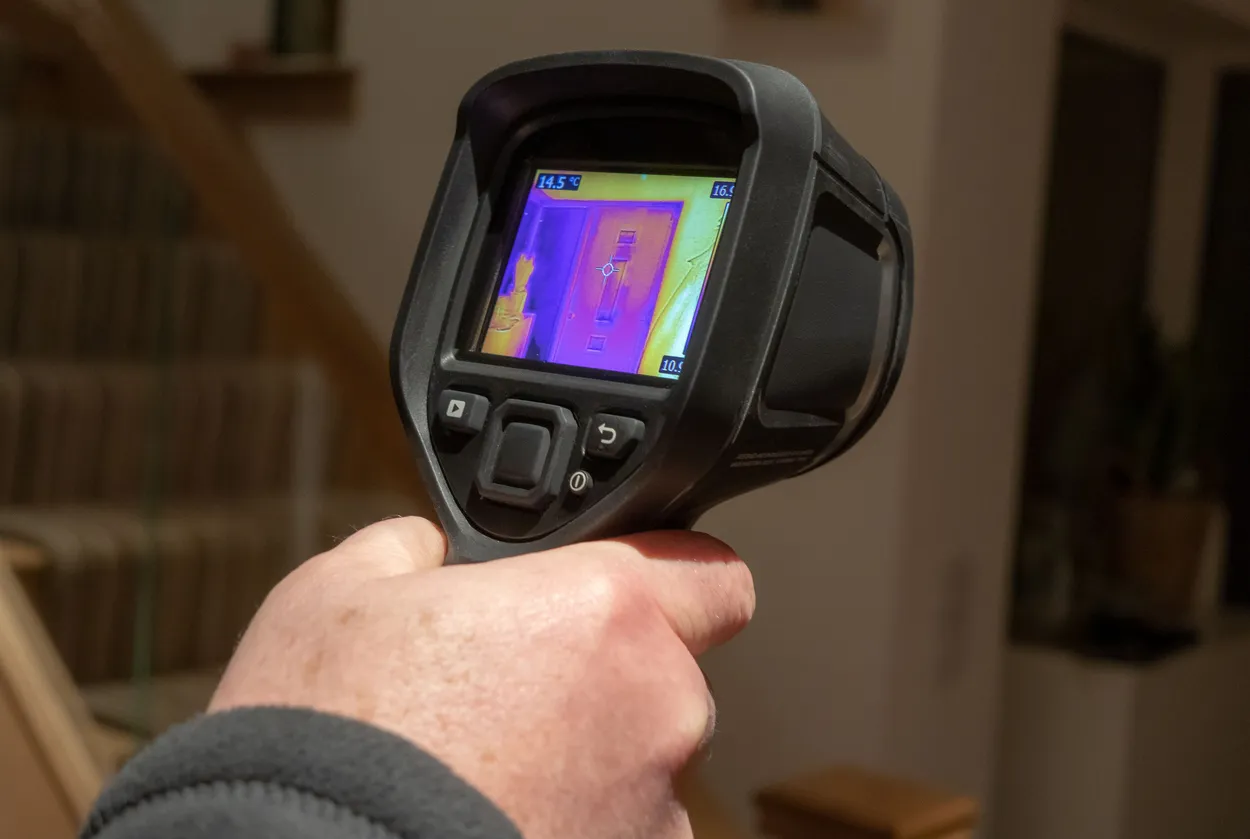 A close-up of a person's hand holding a thermal imaging camera, which displays a colorful heat map of a wall.
