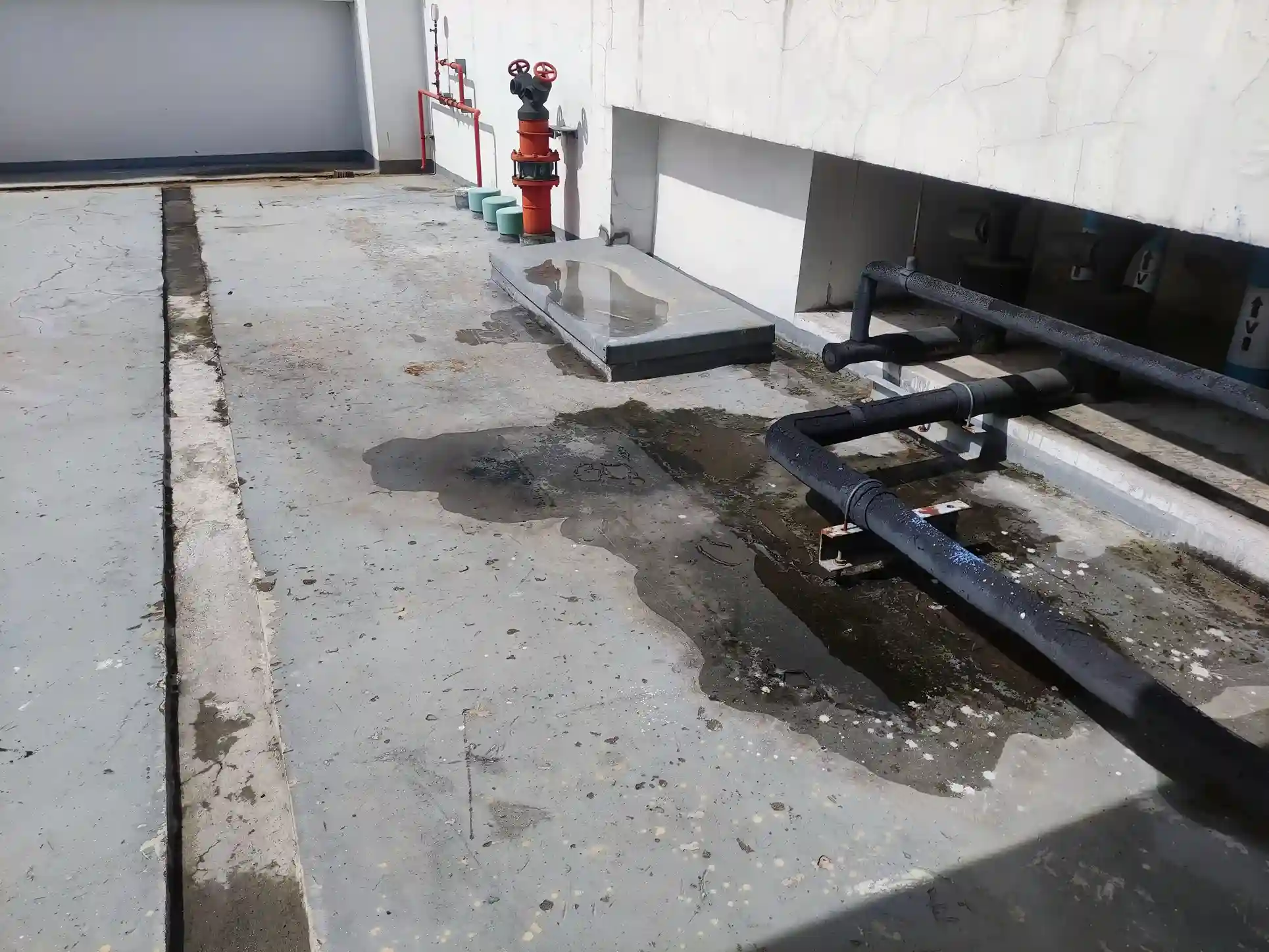 An aerial view of an industrial building's concrete rooftop or foundation, showing a large puddle and wet area around black pipes and a fire hydrant, indicating a slab leak.