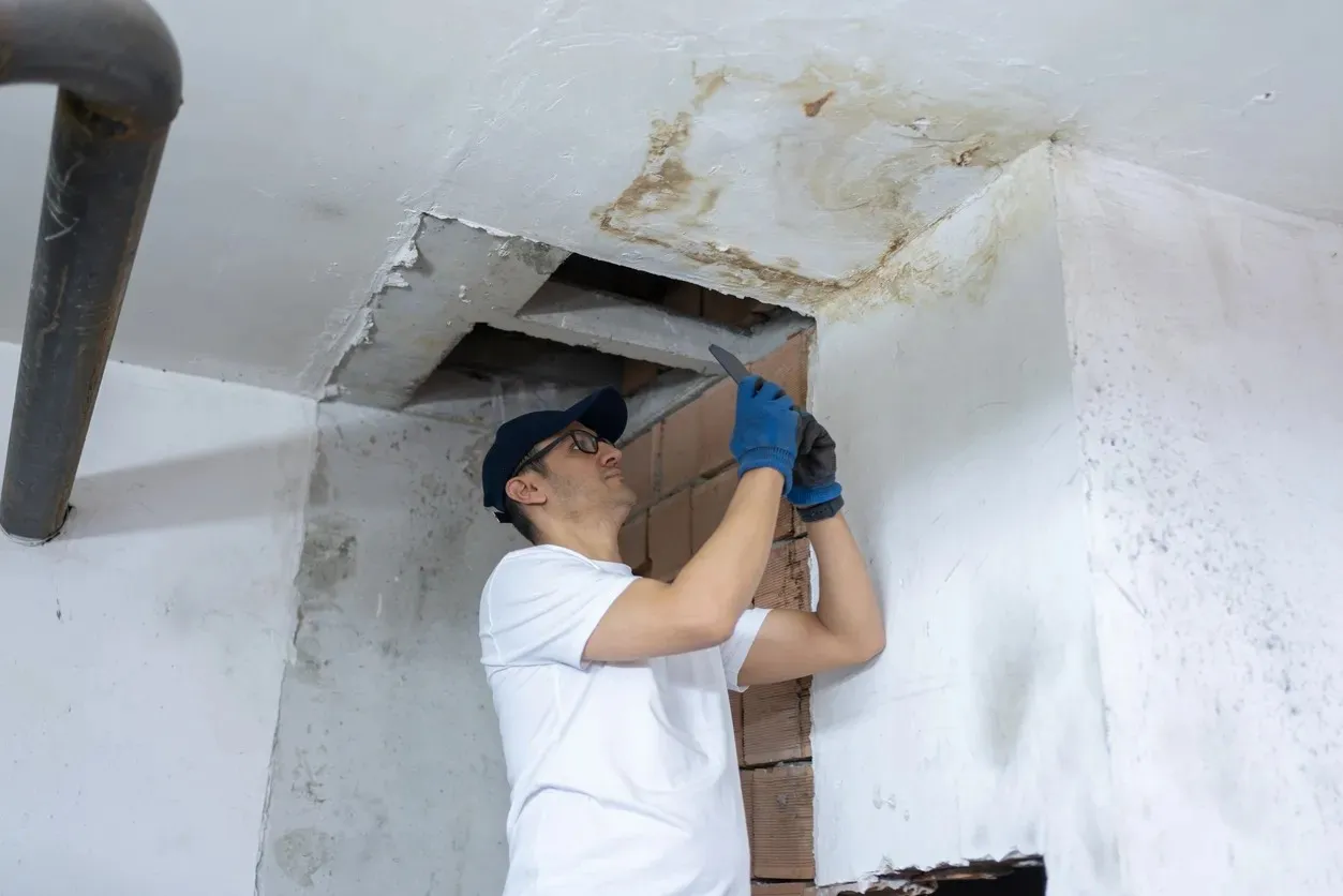 A plumber in a baseball cap and gloves inspects a water-damaged ceiling with a phone or tool.
