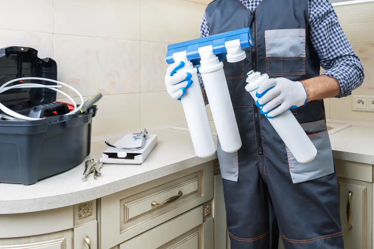 A plumber holds a three-stage water filter system in a kitchen, with a toolbox and various tools on the countertop beside him.