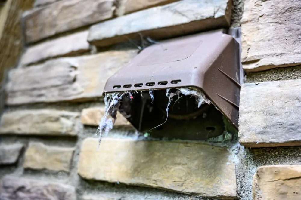 A dirty, lint-filled dryer vent is shown on the exterior brick wall of a house