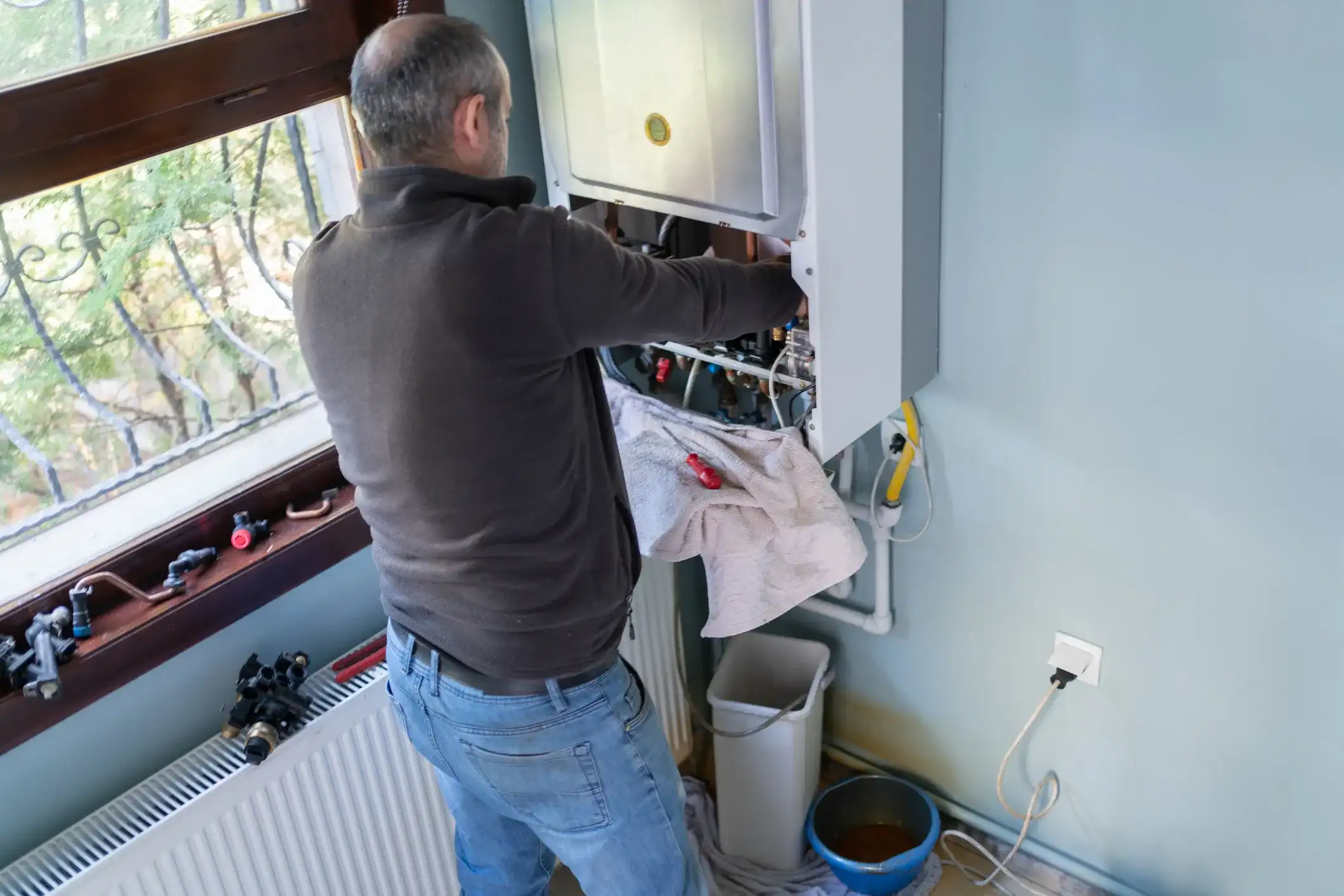 Plumber wearing jeans and a fleece jacket servicing a boiler mounted on a wall.