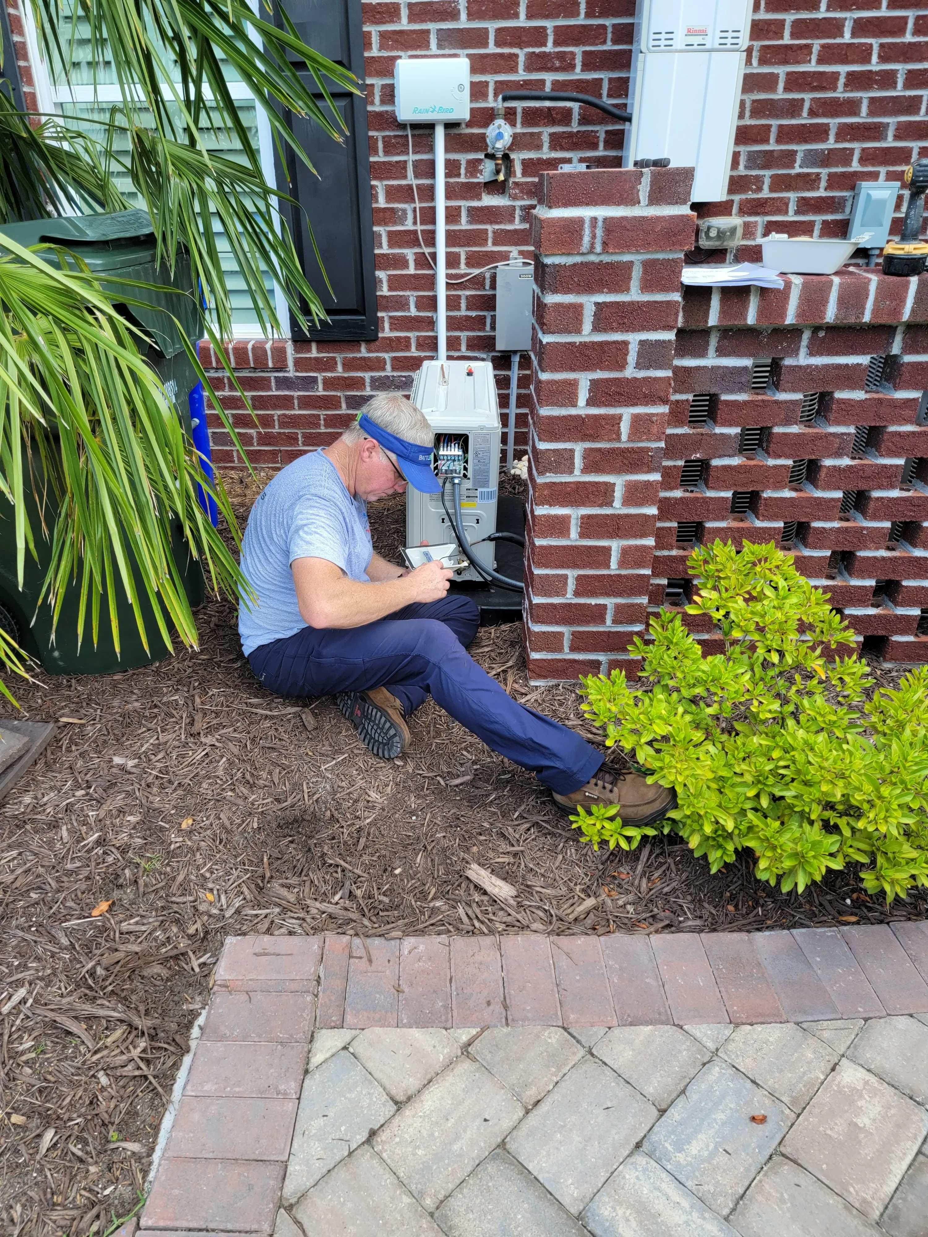 Man in blue cap and shirt works on air conditioner outside a brick building, surrounded by greenery.
