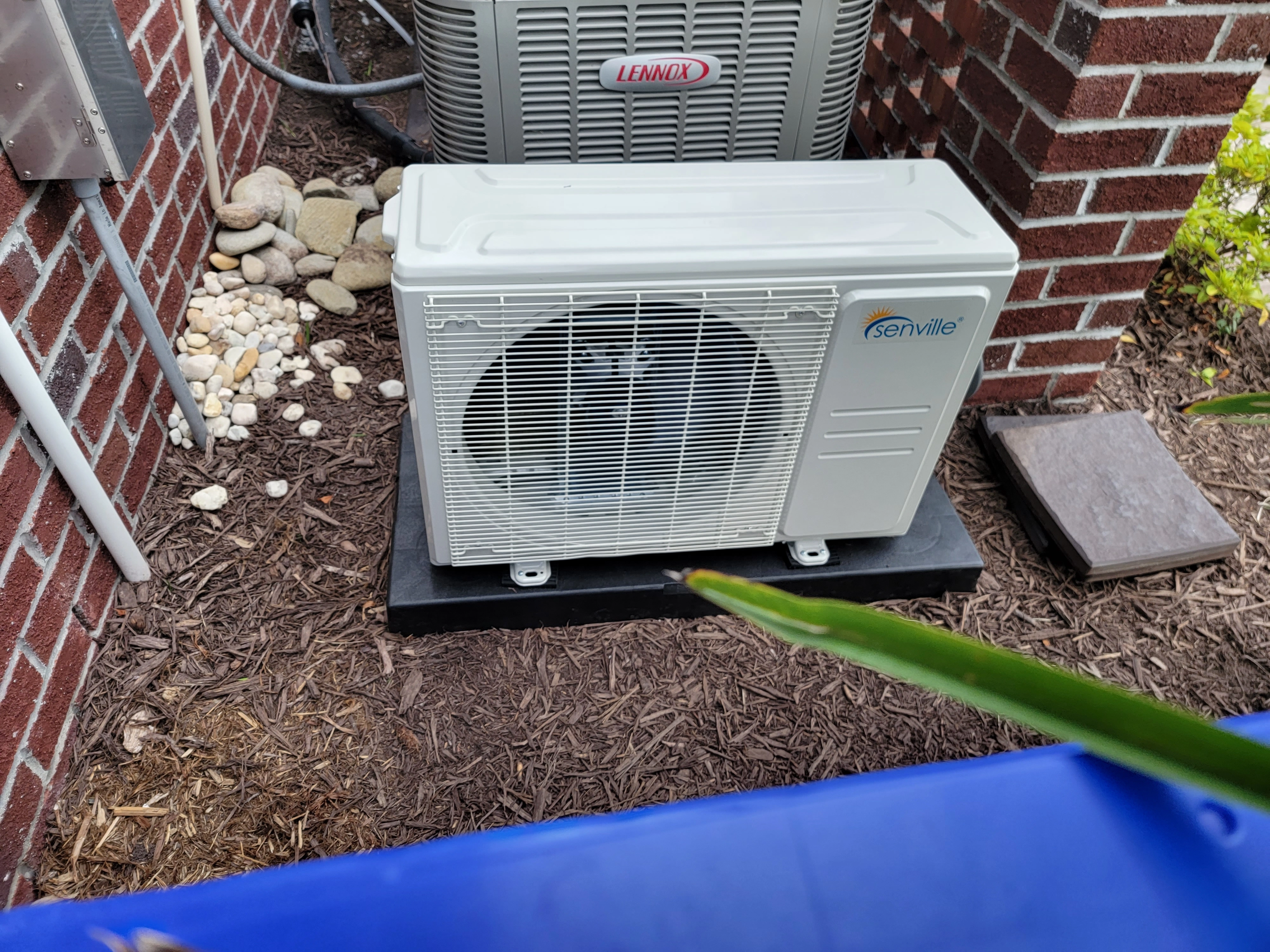 Outdoor air conditioning unit with "Senville" logo on a black base, set against a brick wall.