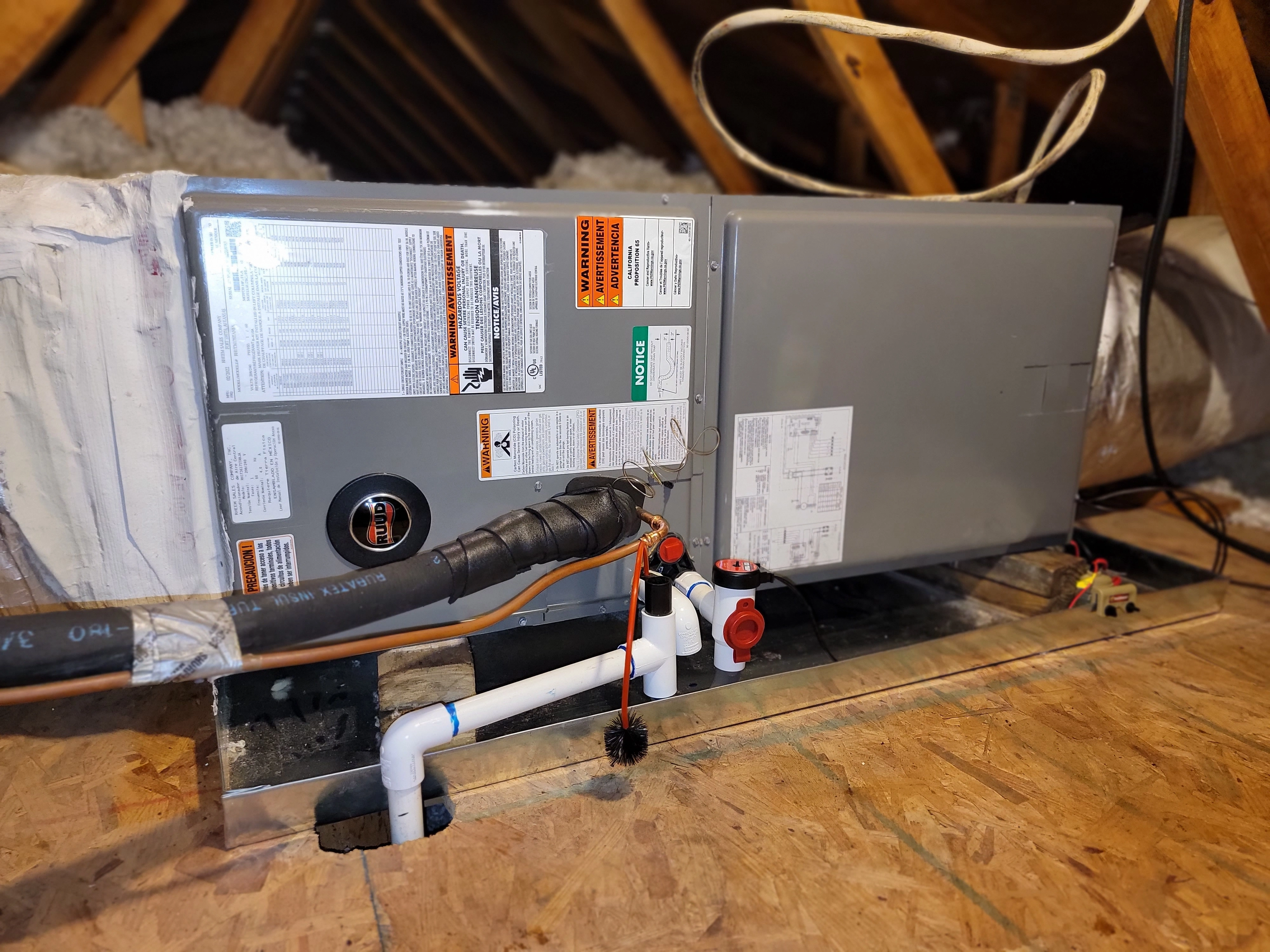 A gray Furnace system, sits on a wooden attic floor with visible pipes and warning labels