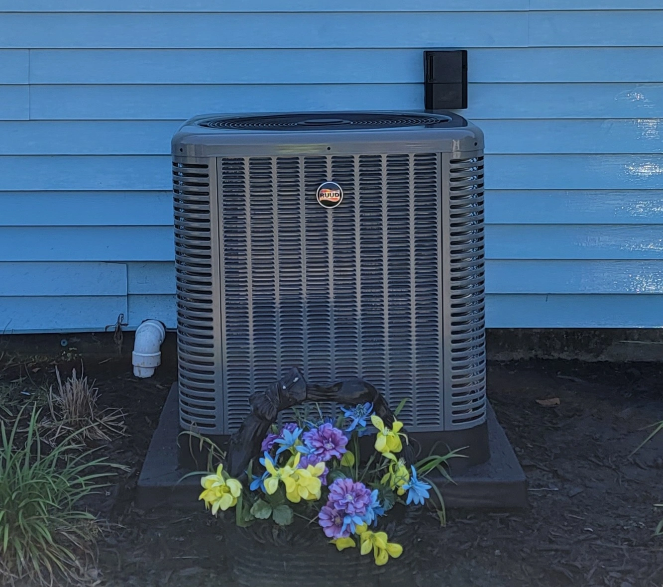 Air conditioning unit against a blue house with a small basket of artificial purple and yellow flowers at its base