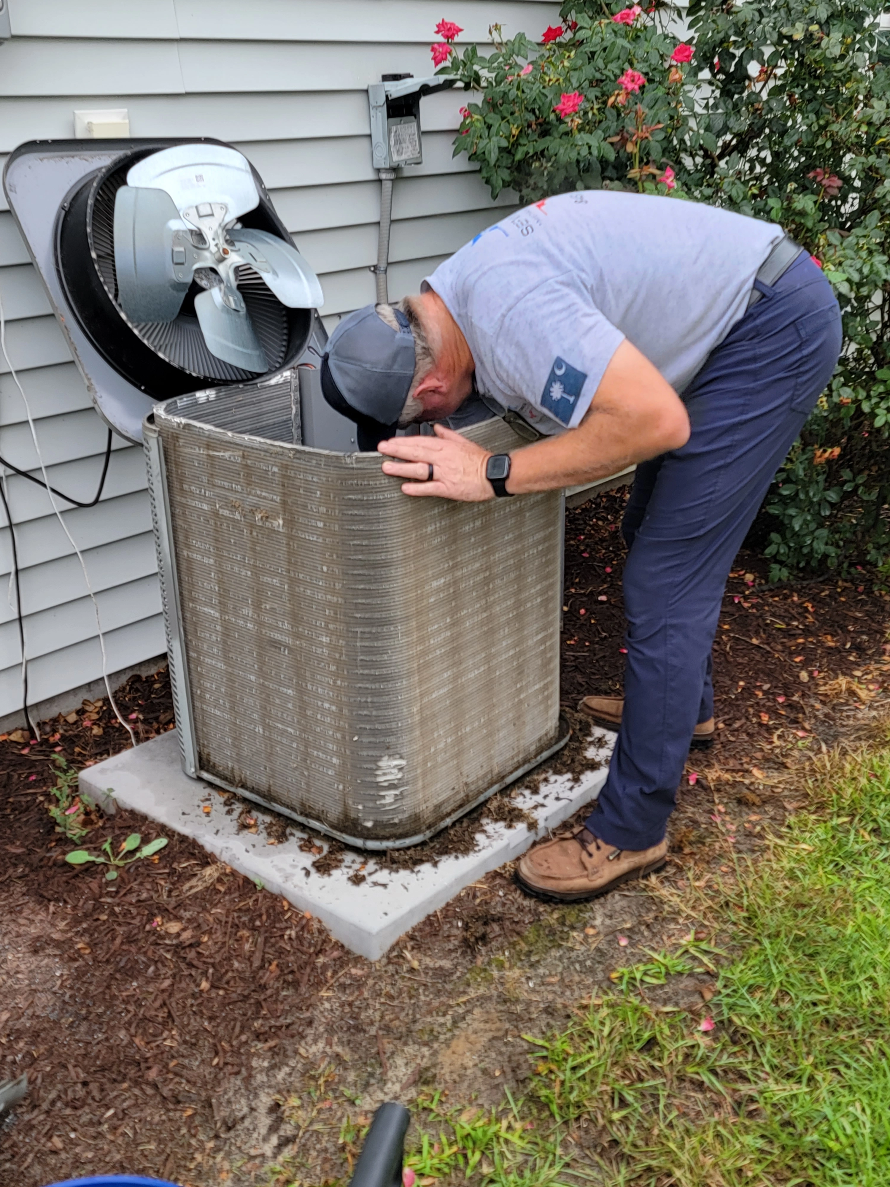 A man in casual work attire inspects an open outdoor air conditioning unit beside a house, surrounded by shrubs
