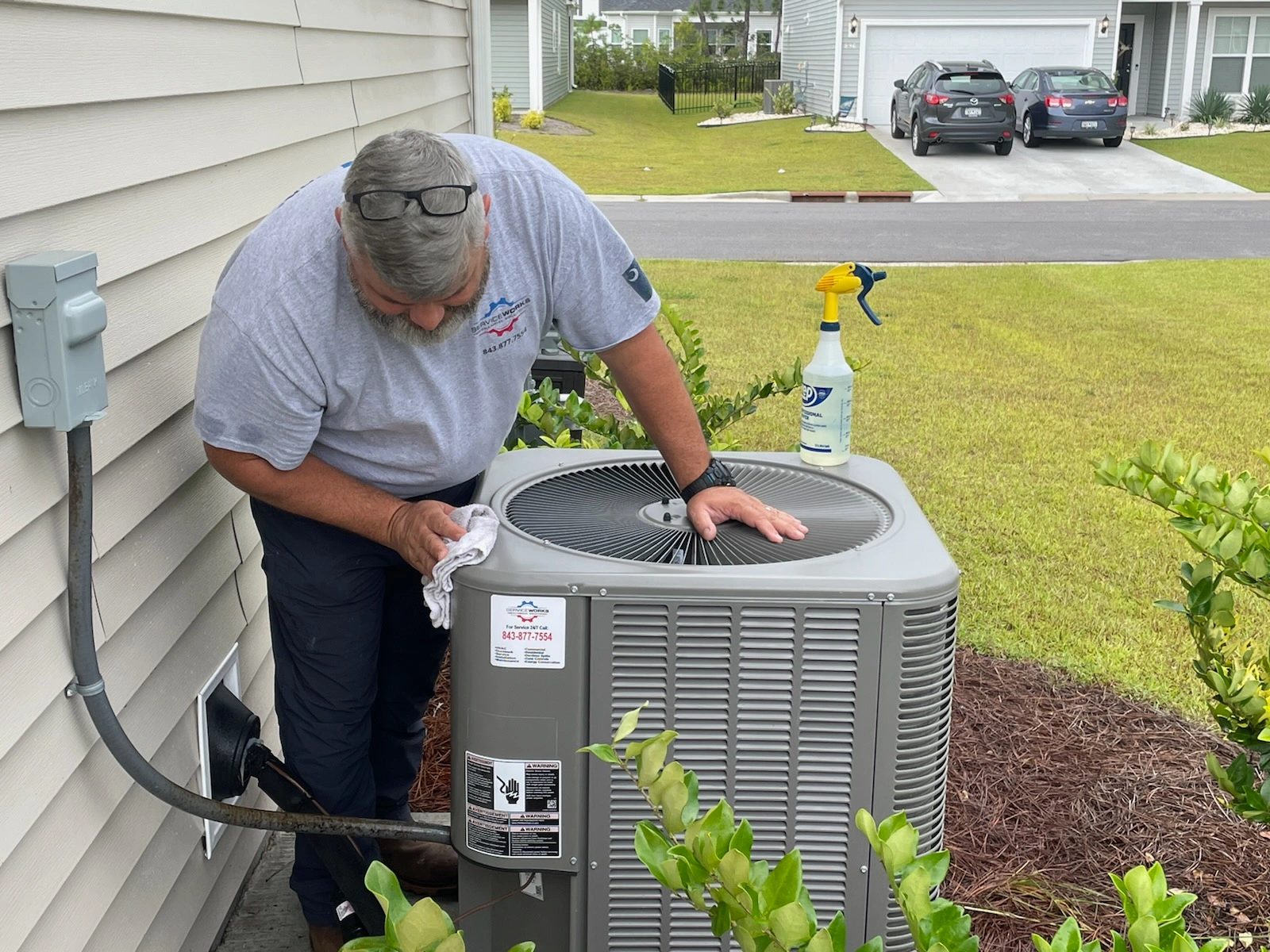 A technician in a gray shirt cleans an outdoor AC unit with a cloth, next to a spray bottle.