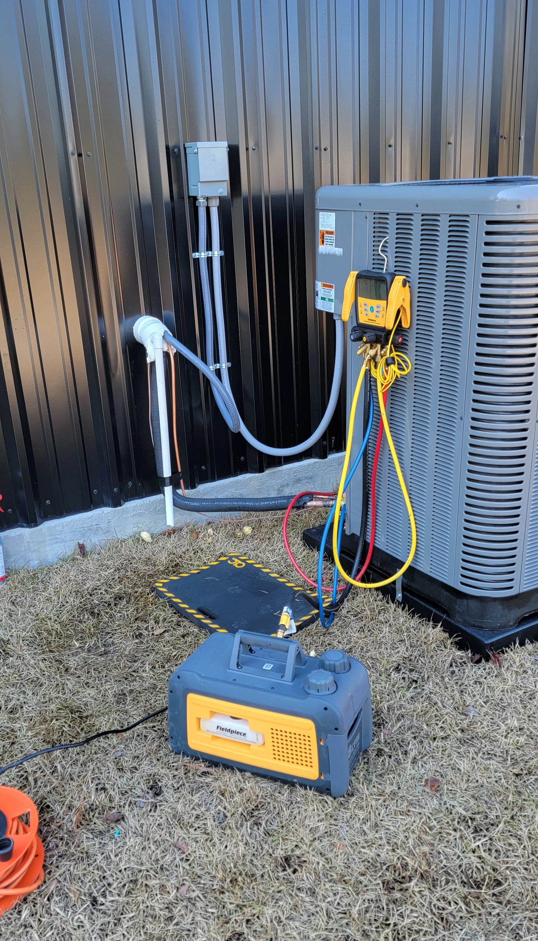 Outdoor air conditioning unit with colored hoses connected, beside a black metal wall.