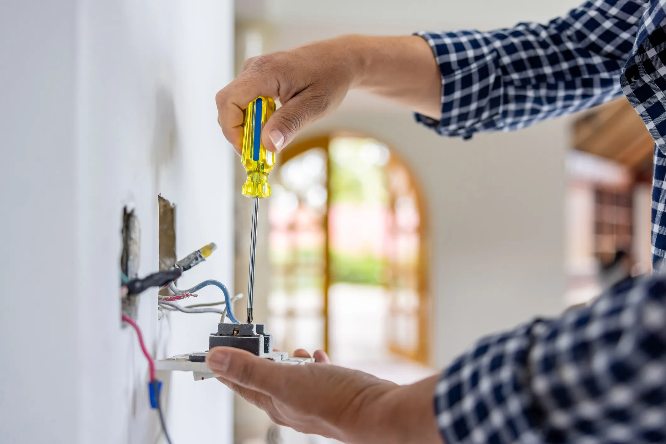 An electrician in a plaid shirt uses a yellow-handled screwdriver to work on a white switch plate with exposed wires.